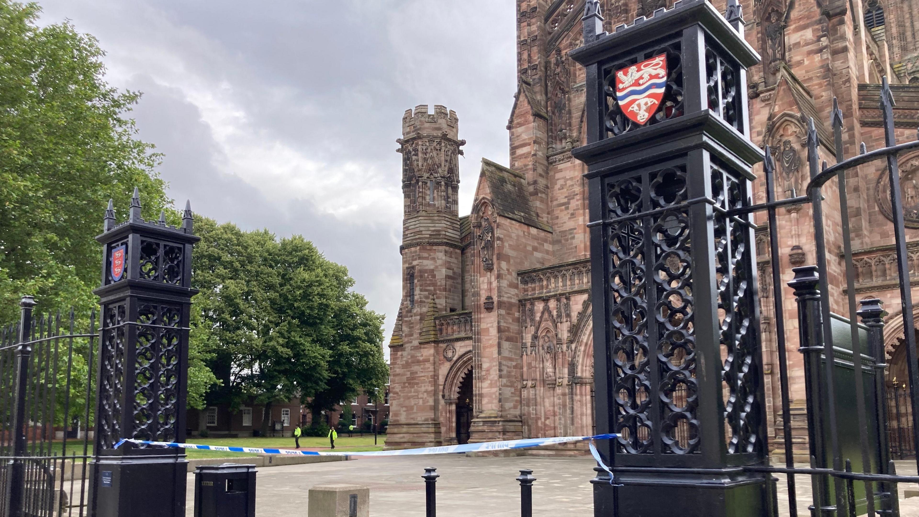 A blue police cordon stretches across the black metal entrance pillars to a large cathedral. The cathedral is in the background, and is made with red-brown bricks. There are green trees and grass to the left of it, and two police officers in yellow hi-vis jackets on the grass. 