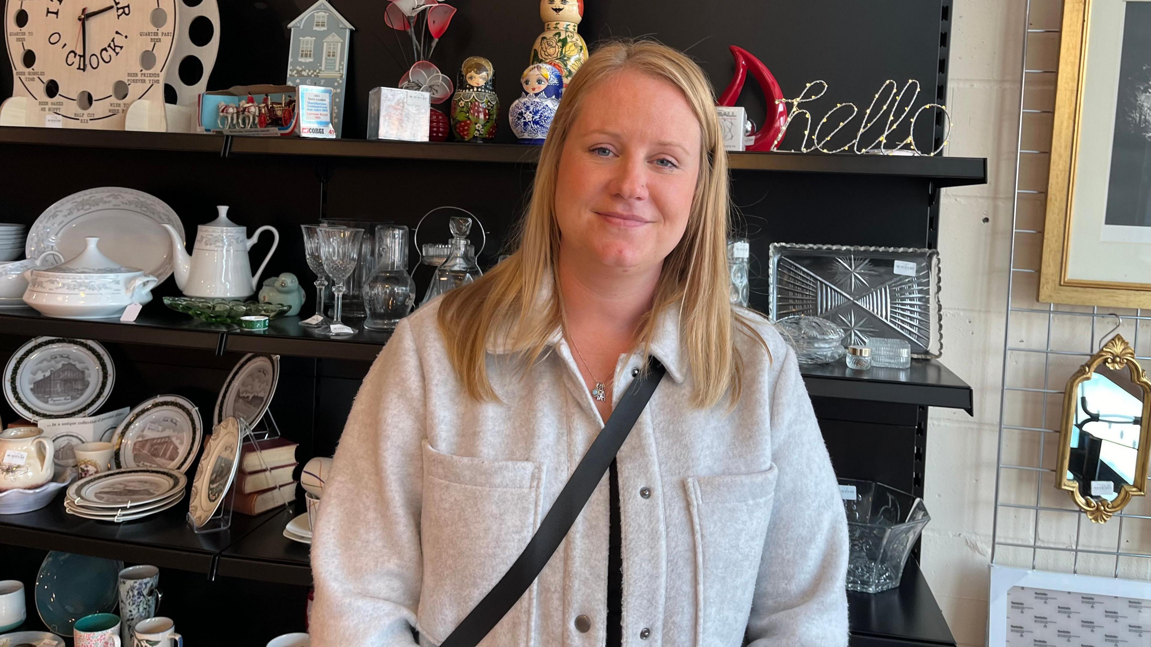 Sam is standing in front of a shelf with crockery. She is wearing a grey jacket and smiling at the camera.