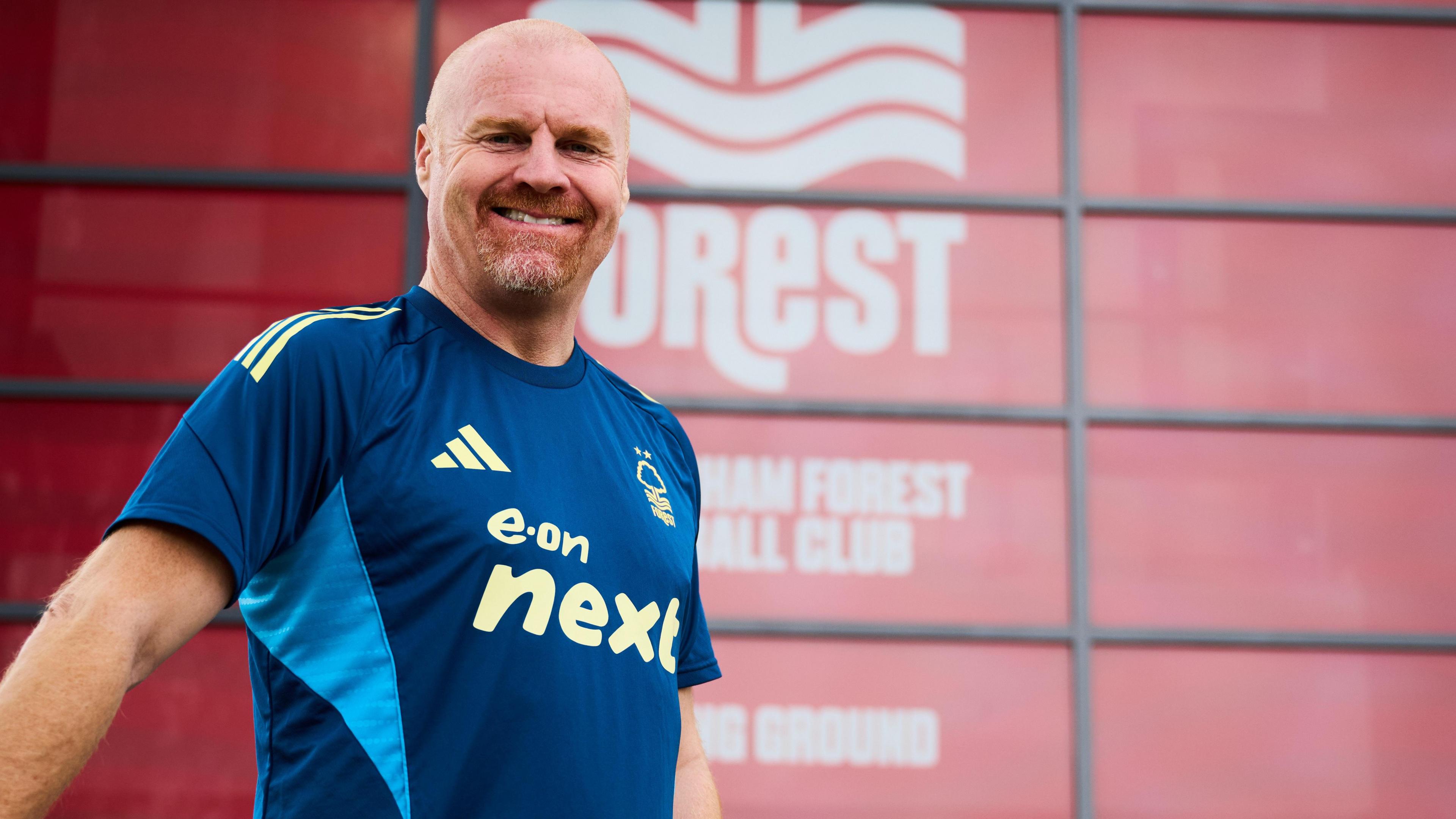 Sean Dyche at Nottingham Forest, wearing a blue football top, standing by a wall, smiling and looking at the camera. He has a red bear and bald head.