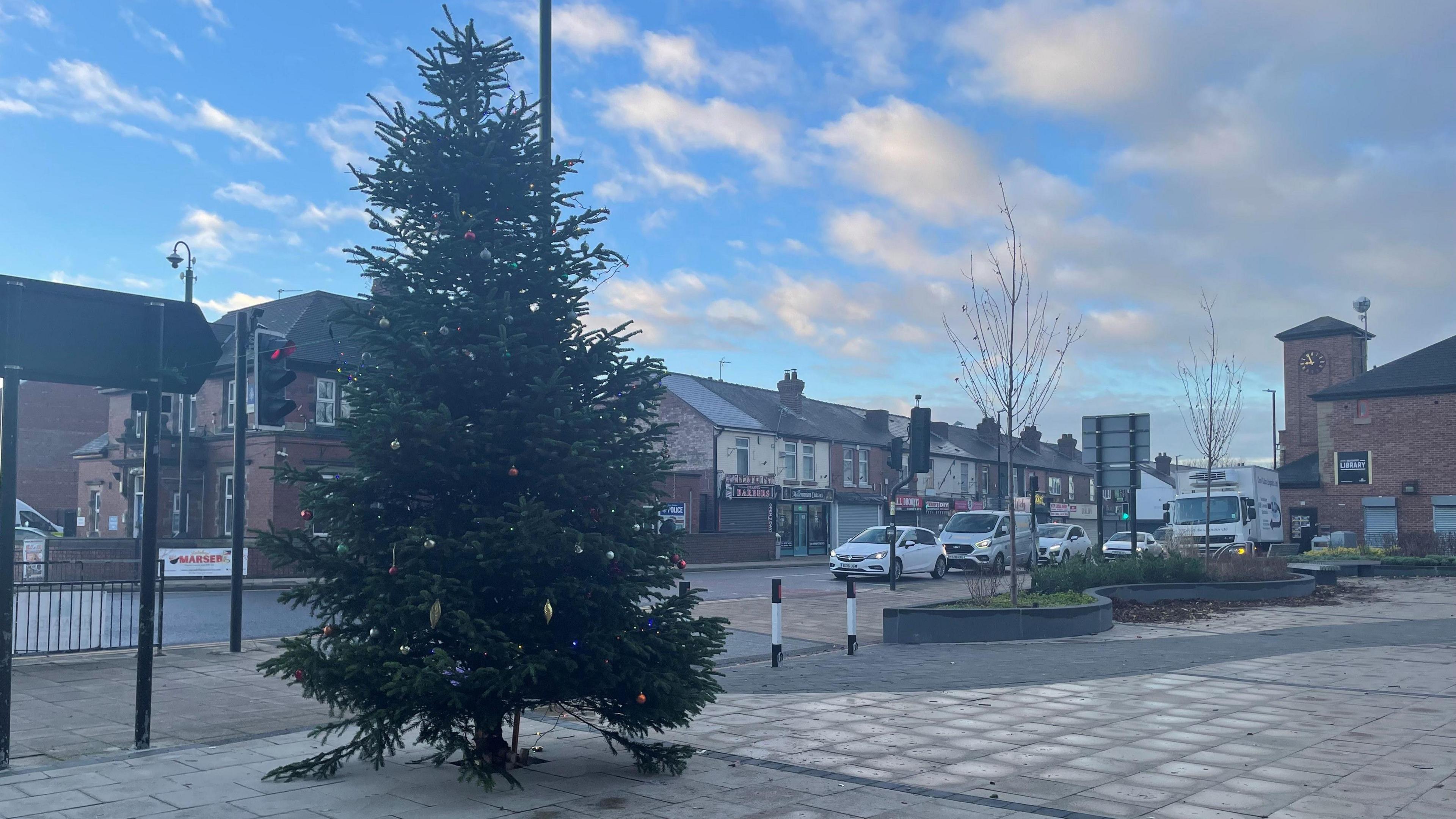 Christmas tree in a town centre. A road in the background has traffic held up at traffic lights. 