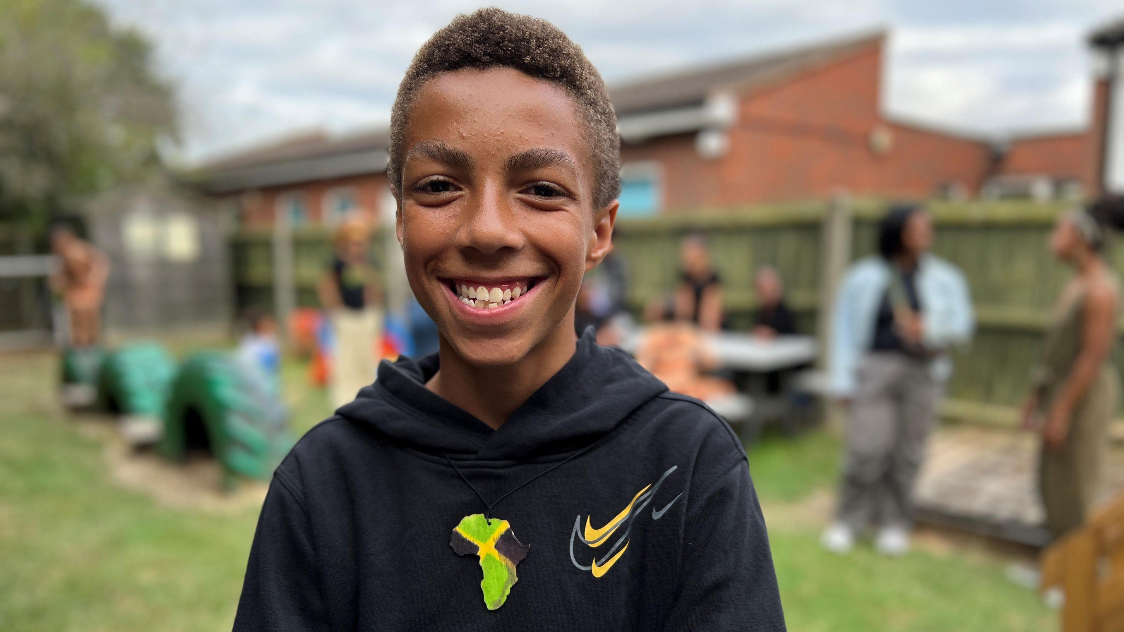 Ricardo smiling at the camera wearing a black hoodie. The background with other kids at the school has been blurred