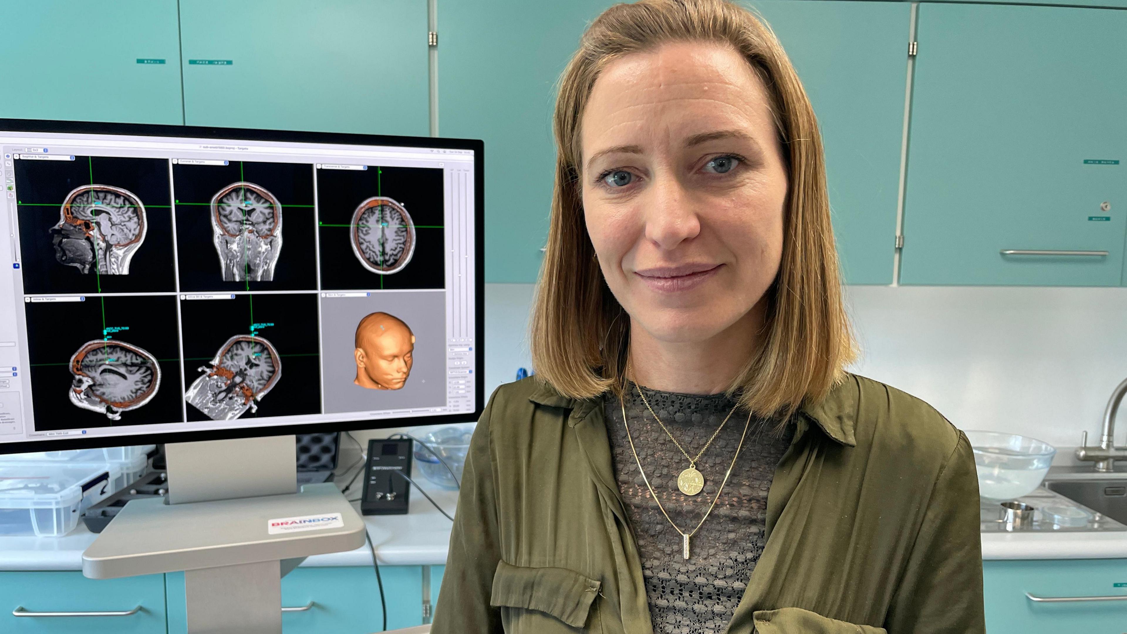 A woman with bobbed blonde hair, wearing casual green clothes. She is standing in front of a computer screen showing scans of parts of a brain 