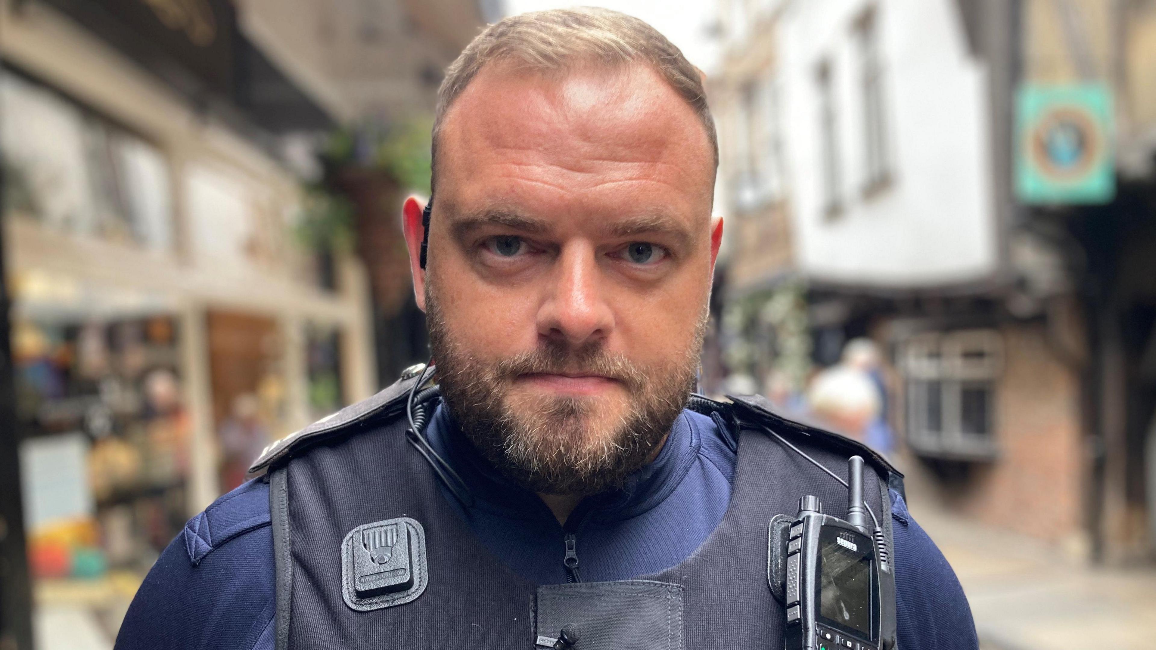 Immigration officer Richard Garbutt is stood on a street in York. He has short brown hair and beard and is wearing dark blue clothing with a black vest with a radio on on left shoulder