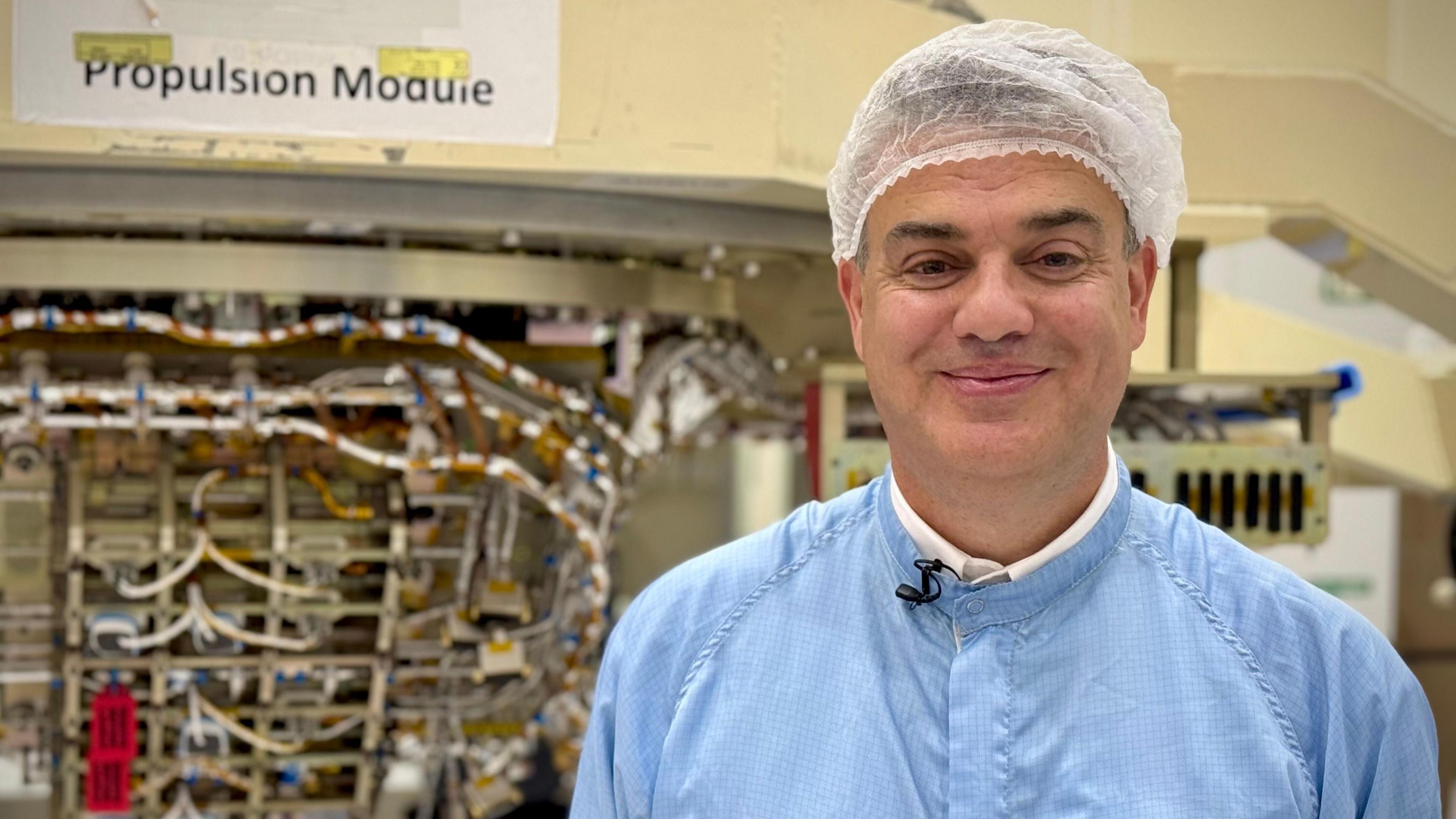 A smiling man wearing a hair net and dressed in a blue science coat is stood in front of cables labelled "propulsion module"