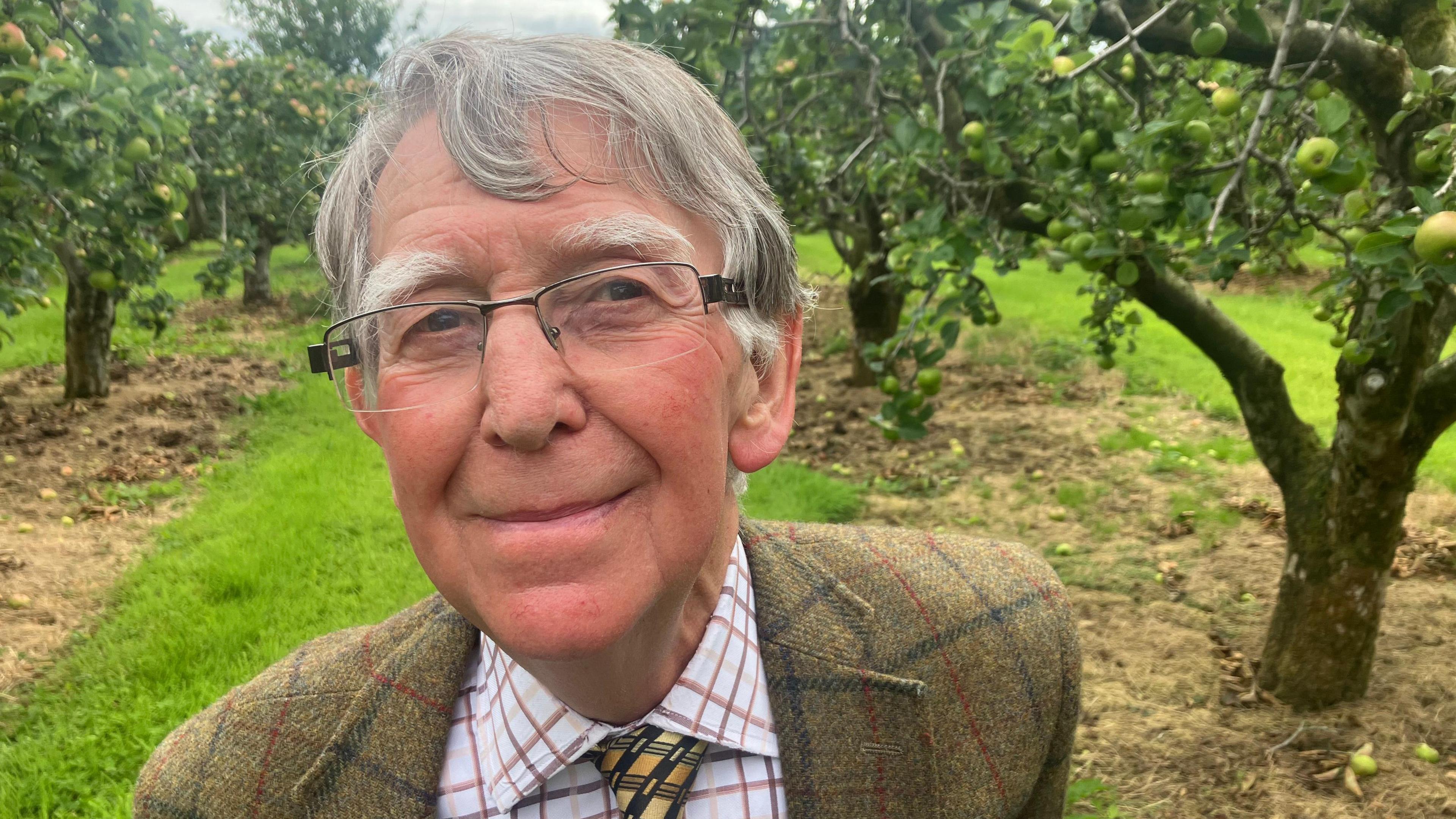 Man in green checked blazer with yellow and black tie. Grey hair and wearing glasses, in front of apple tree.