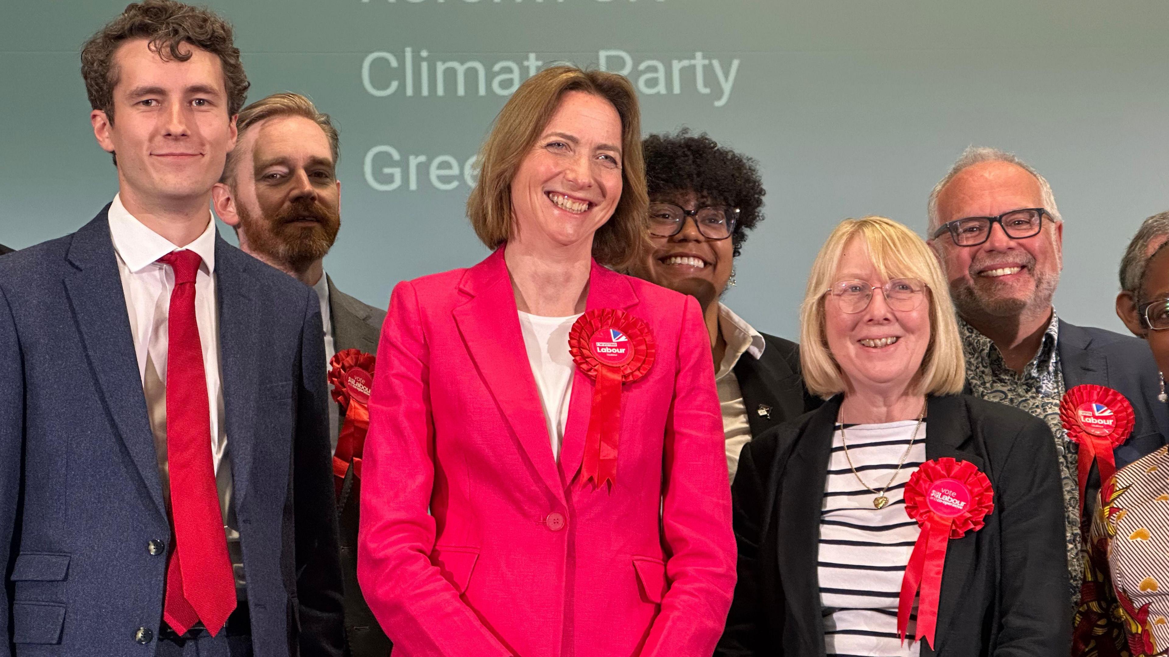 Six people standing on a stage and smiling. In the centre is Colchester MP Pam Cox, who is wearing a pink suit and a red rosette. She is standing with members of Colchester's Labour Party.