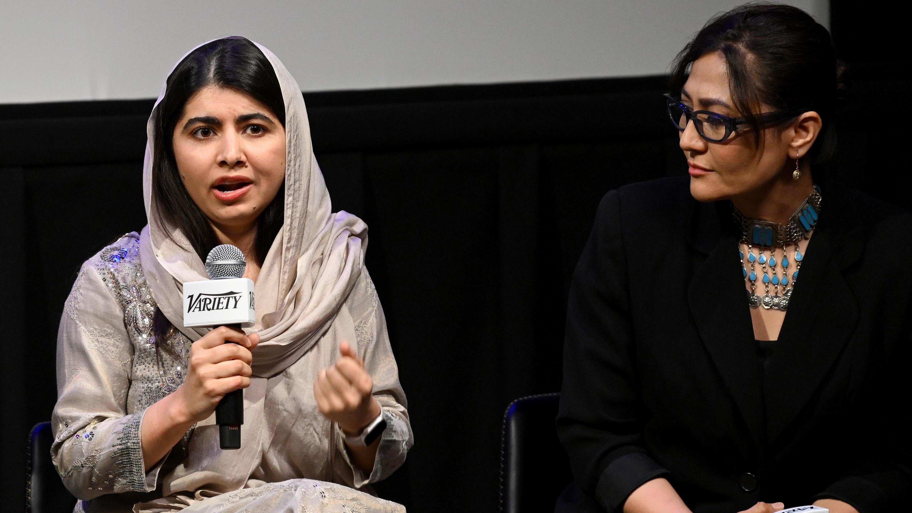 Malala Yousafzai and Sahra Mani on stage at an event in New York. Malala holds a microphone as she addresses the audience. She wears a pale grey dress and headscarf, embellished with gems. Sahra sits to her left and looks at her as she speaks. Sahra wears a black top and blazer with an elaborate blue necklace. 