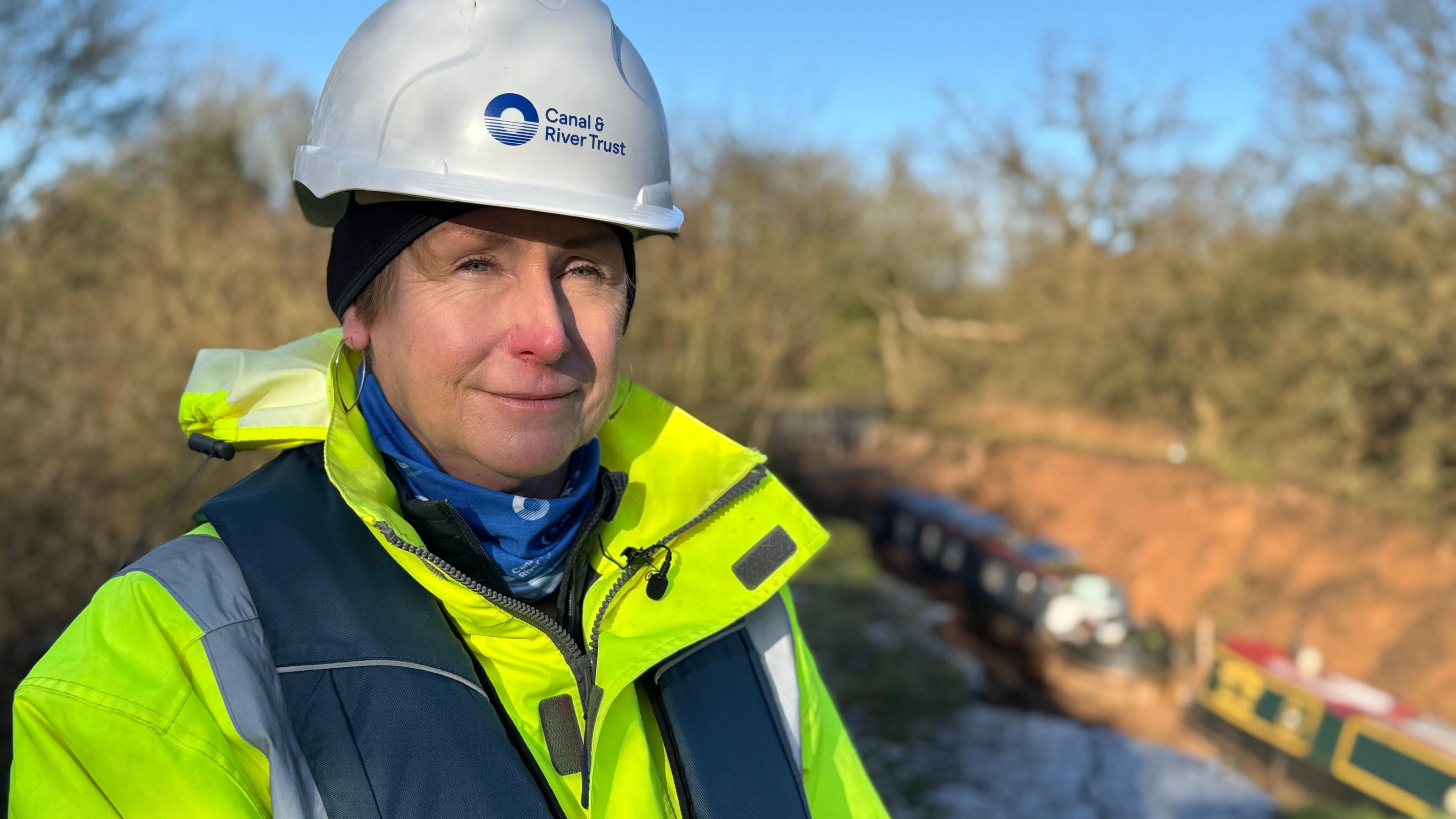 A woman in a white hard-hat which says Canal & River Trust on it. She wears a hi-viz jacket and stands in front of the site of the canal embankment collapse in Shropshire. Two boats can be seen in the hole left by the incident.