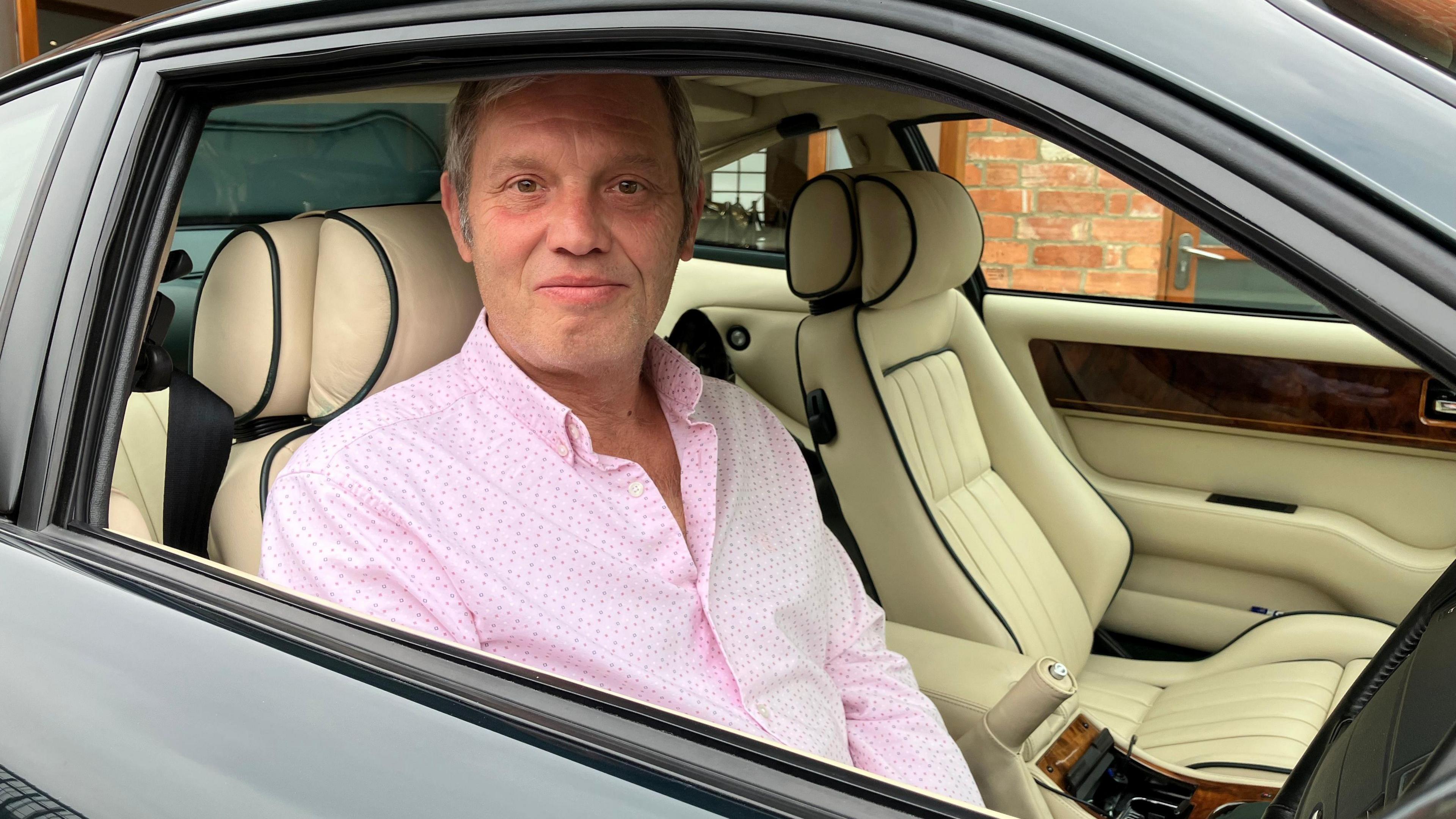 Roger James can be seen sitting in the drivers seat of his car, with the front window down. He is looking at the camera and smiling. Part of the car can be seen. It is green and the seats are cream and leather. The passenger seat is empty.