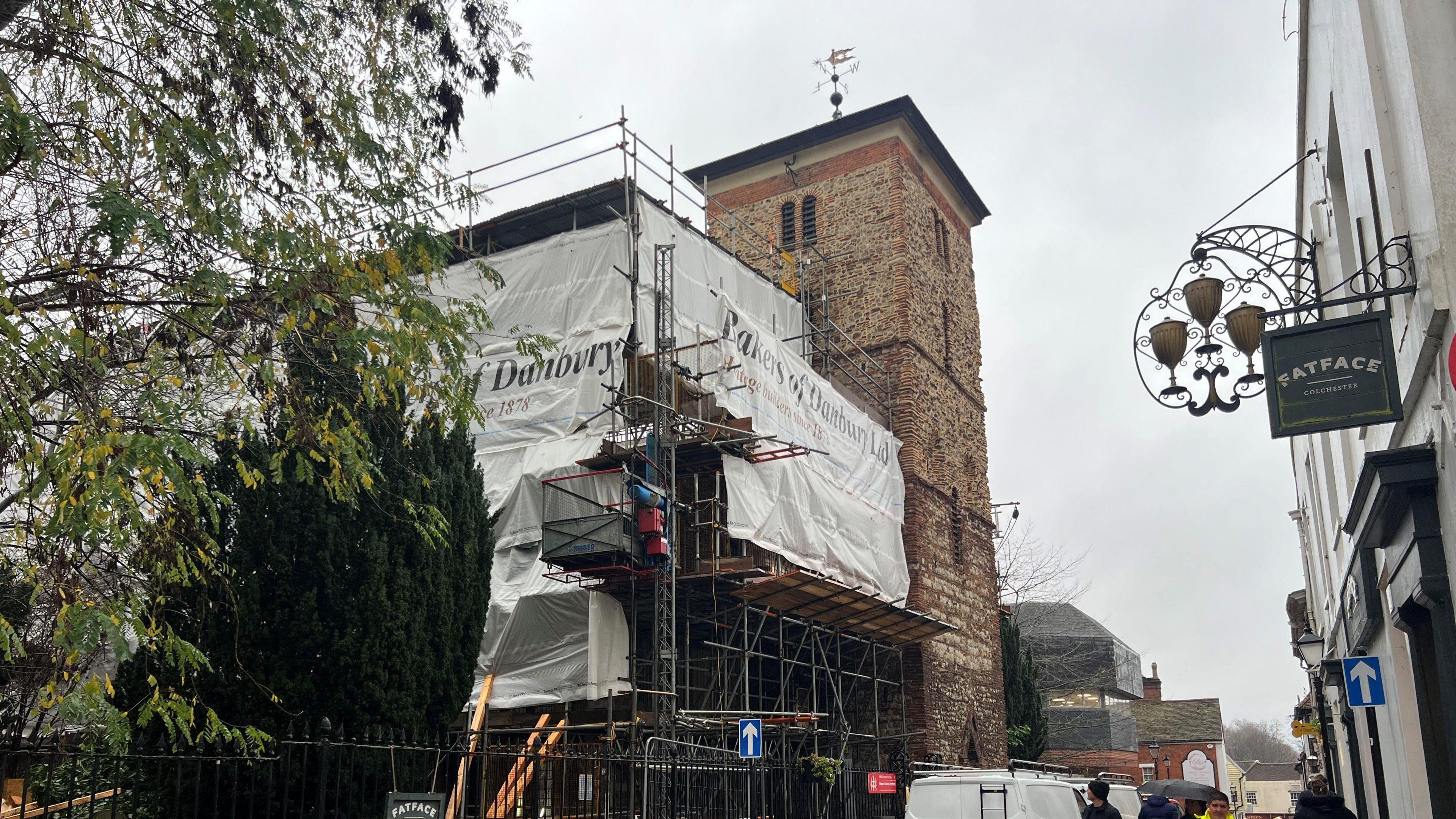 A church covered in scaffolding on the left of the picture. A white curtain has been around the scaffolding. On the right side, in the foreground, are shops, including FatFace.