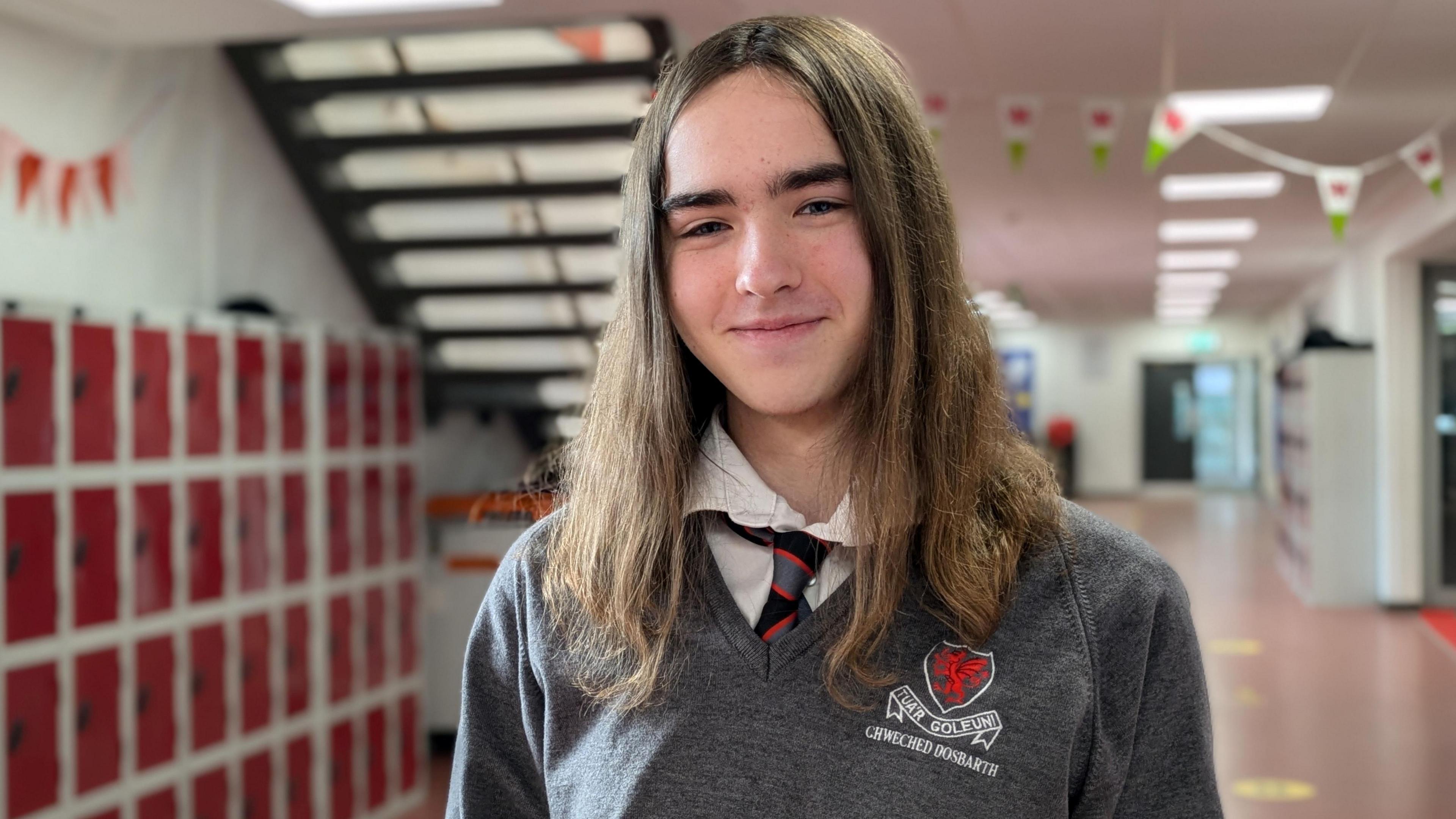 Alexia smiles into the camera. She is wearing a grey school jumper and has long brown hair. She is standing in a school hallway, with red lockers, stairs and Welsh flag bunting in the background. 