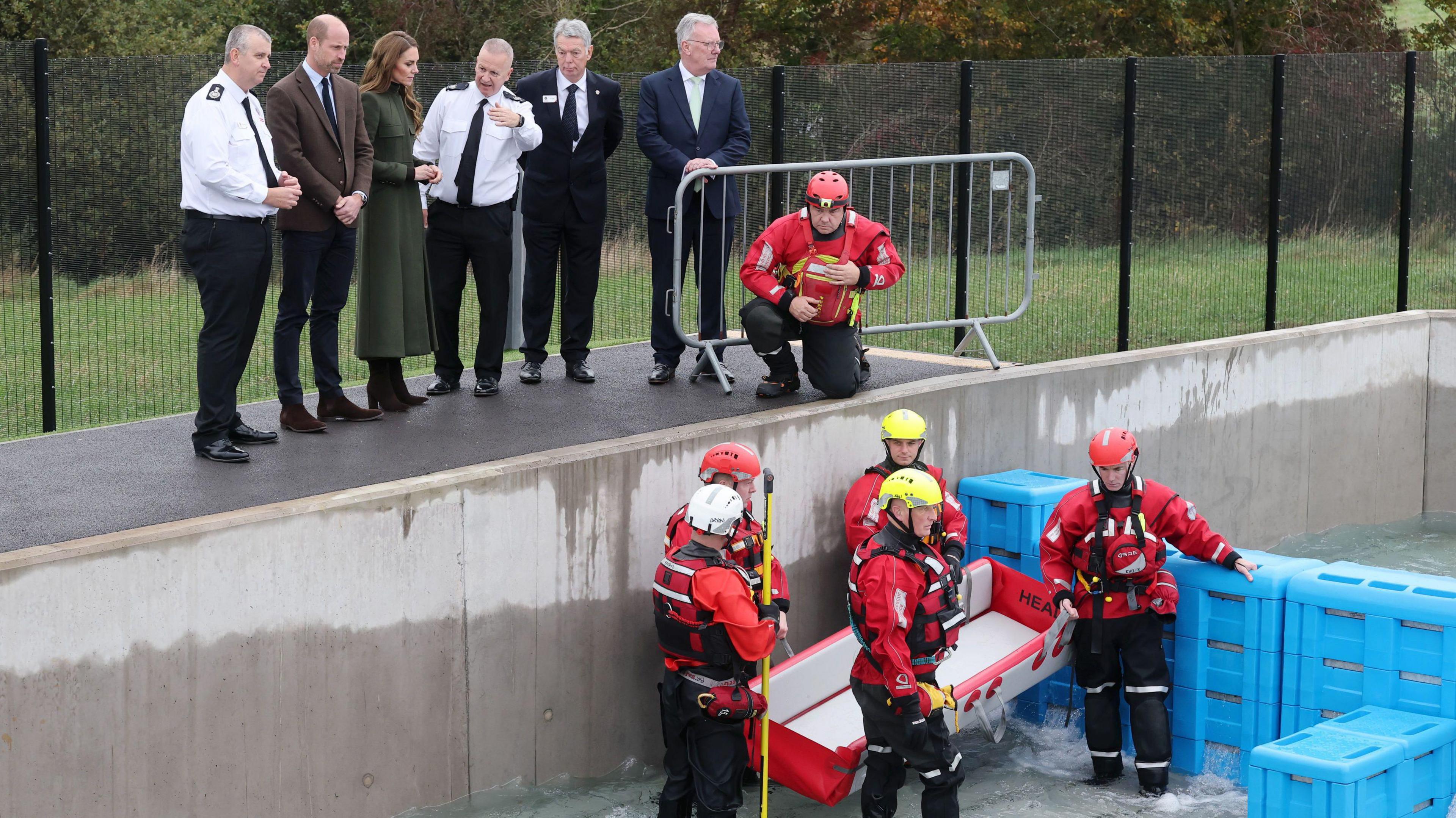 The Prince and Princess of Wales observe a training scenario during a visit to the Northern Ireland Fire and Rescue Service Learning and Development College near Cookstown. They are standing on a platform with a group of men above a deep training pool. Five firefighters in red and black rescue uniforms are standing in the pool holding a stretcher as the level of water increases.