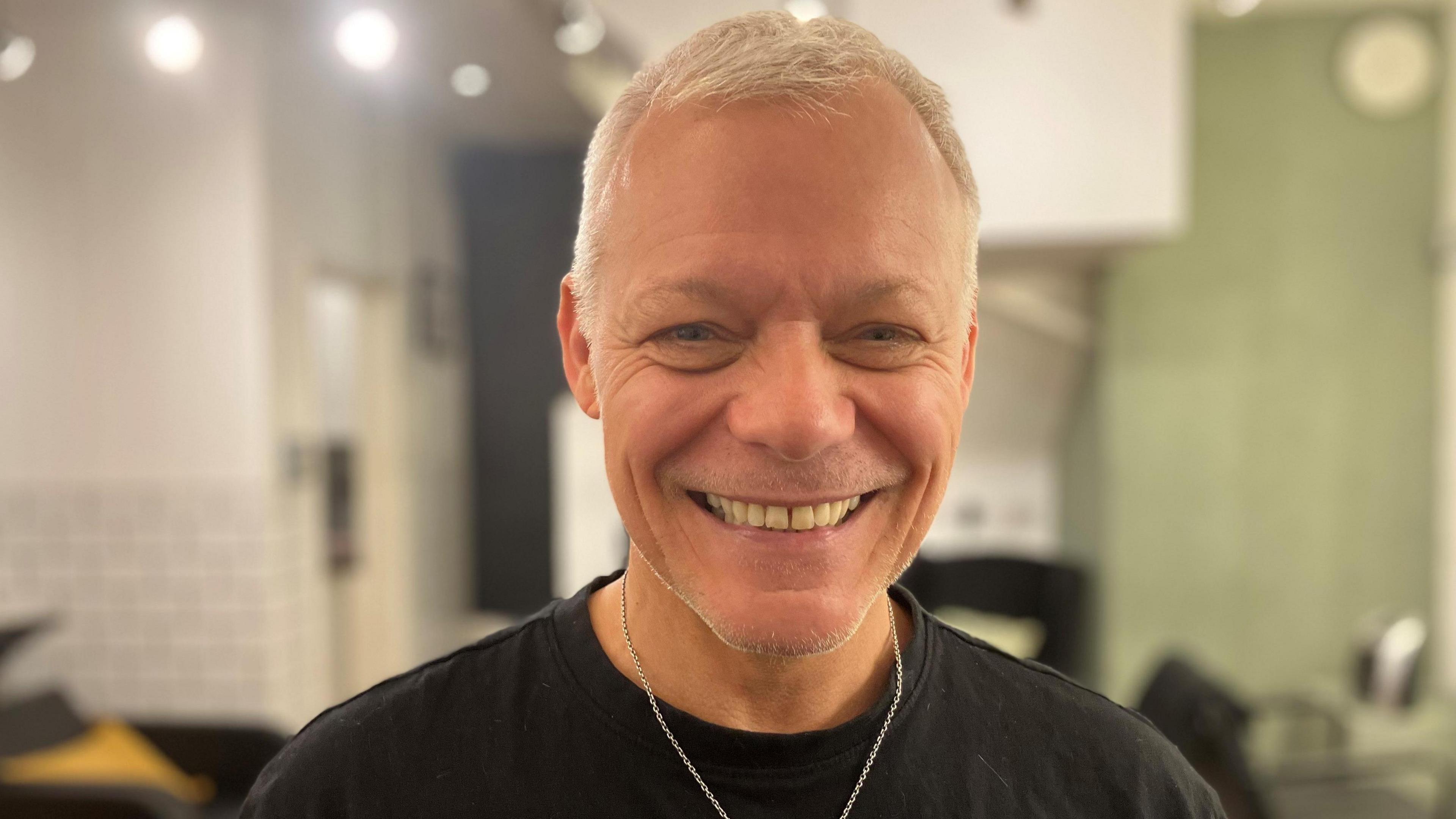 Craig Chapman, who has short blond hair and is wearing a black T-shirt with a silver chain around his neck. He is smiling at the camera.