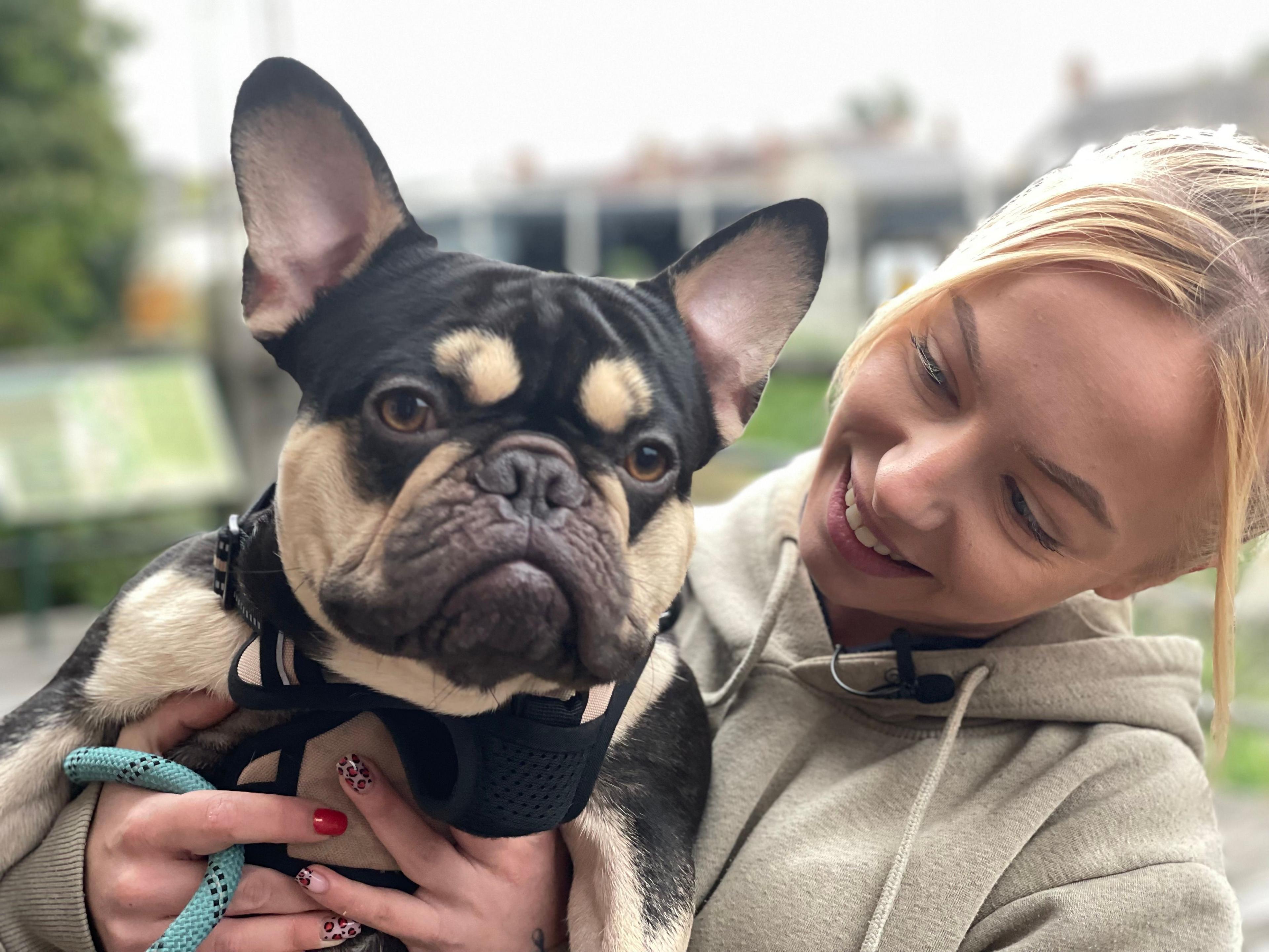 French bull dog with poiunty ears is being held up to the camera by owner Alice who smiles at him.