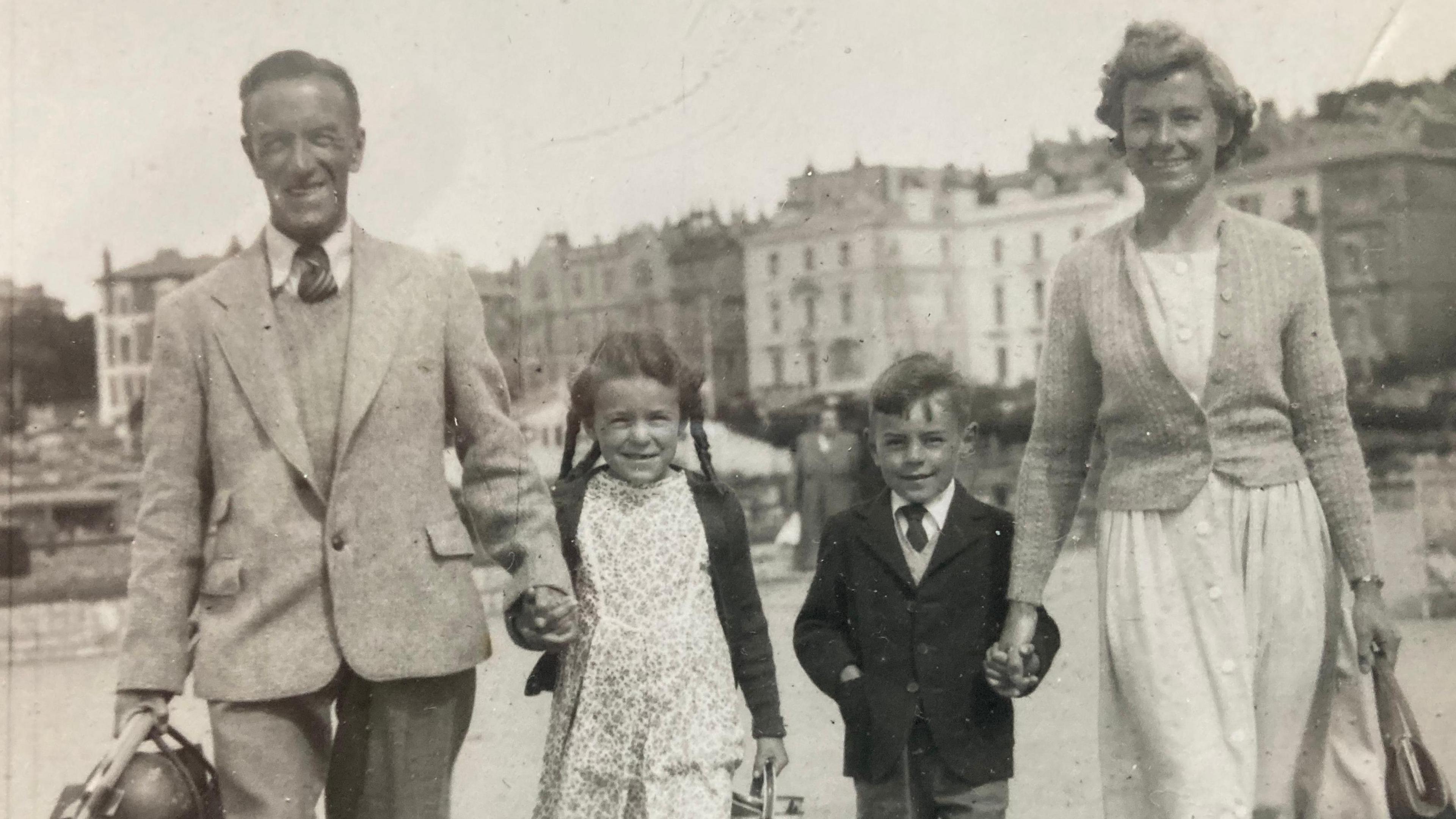 Black and white photo of a family of four at the beach. The mother is on the right and holding hands with the son. Father is on the left holding hands with the daughter. 