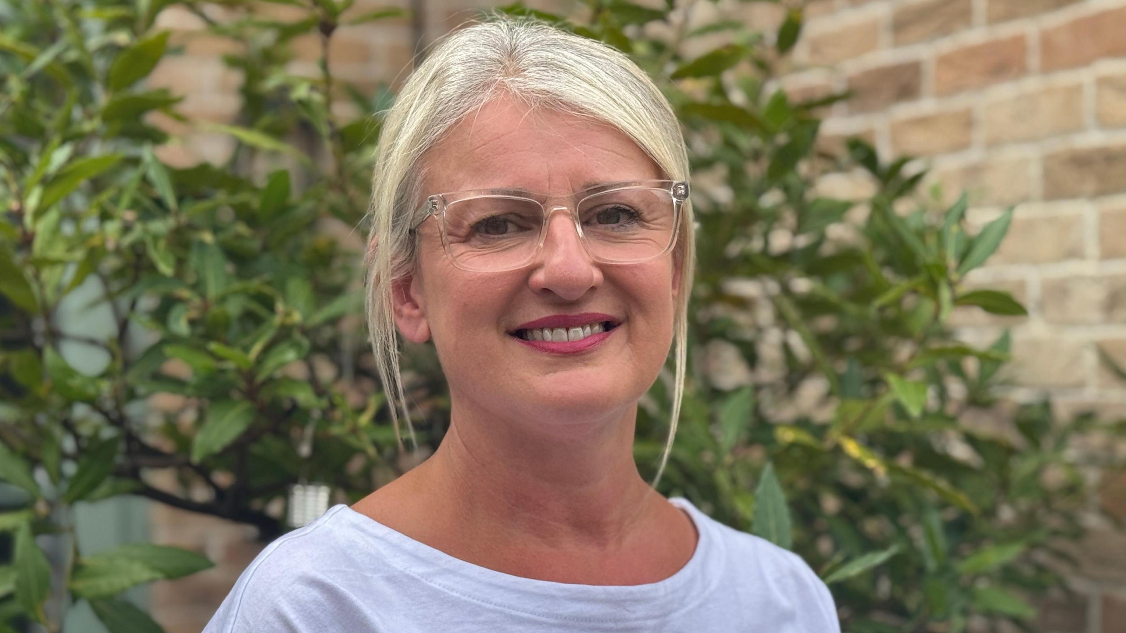 Annys Bossom is standing in her garden smiling at the camera. She is wearing clear-framed glasses, a white T-shirt and her blonde hair is tied back in a ponytail.