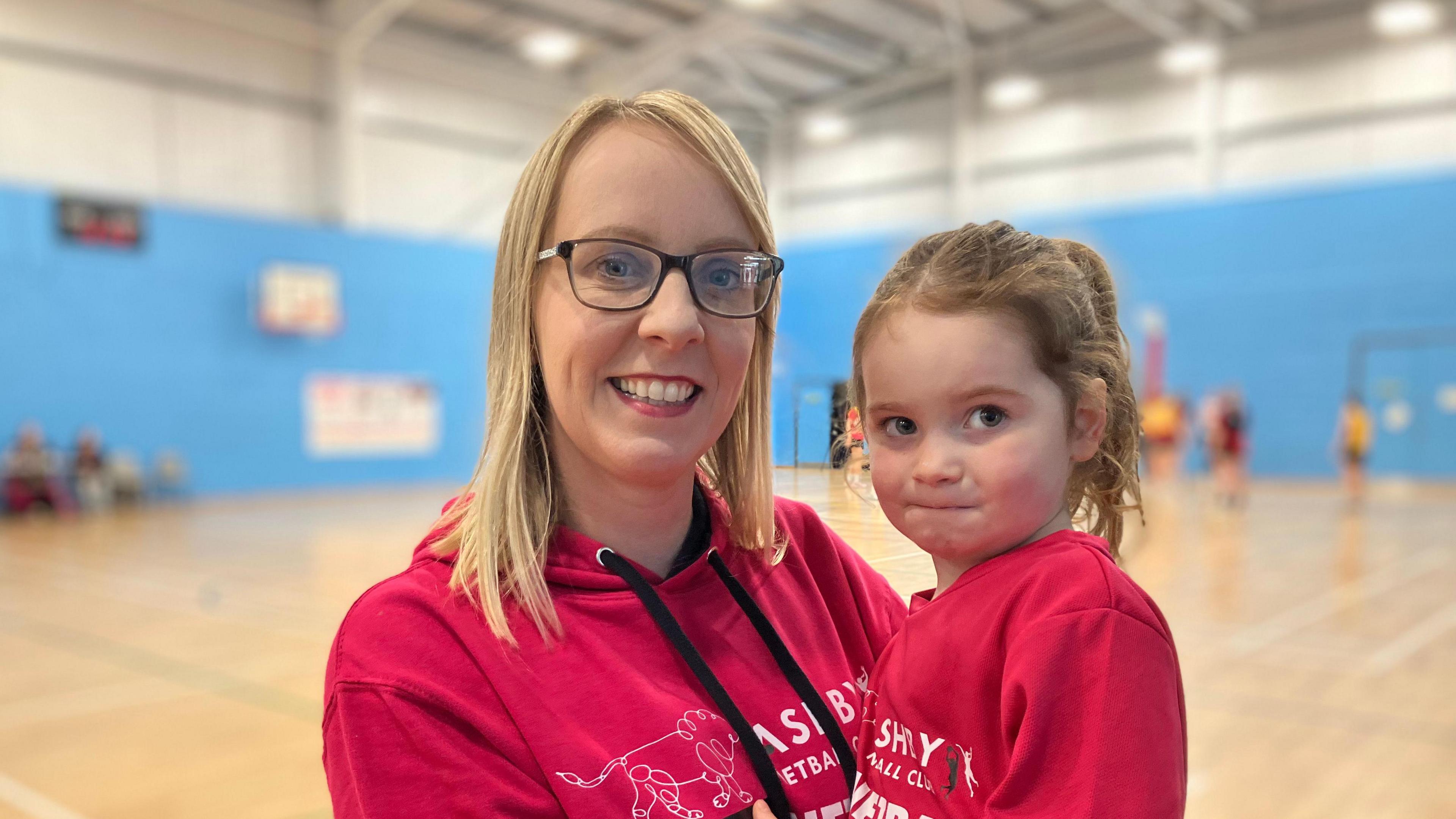 A woman with blonde hair and glasses, holds a little girl with brown hair.  Both wear red shirts and are on a netball court