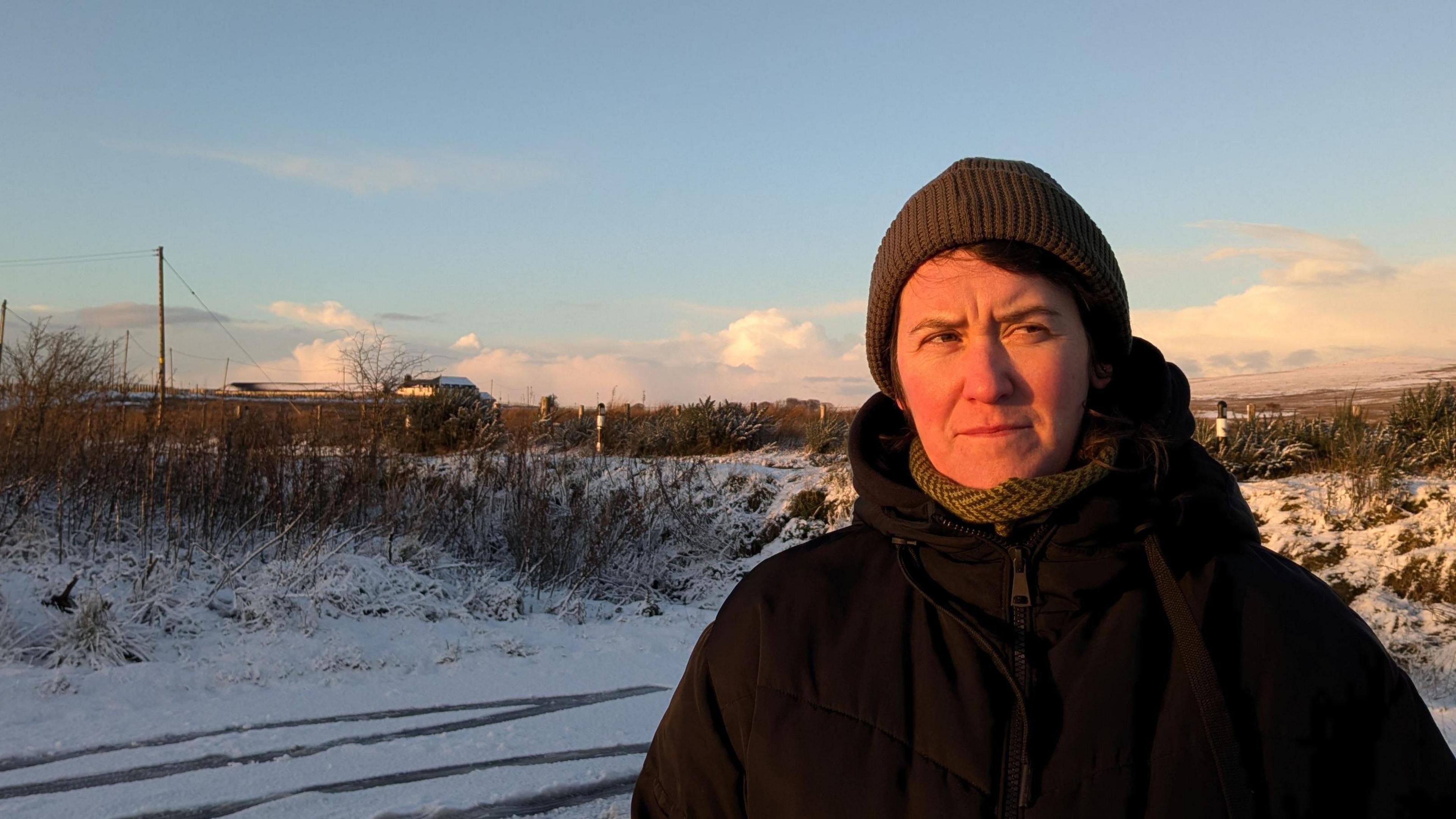 a woman in a beanie hat and thick puffy coat standing in a snowy area, with golden evening light on her face