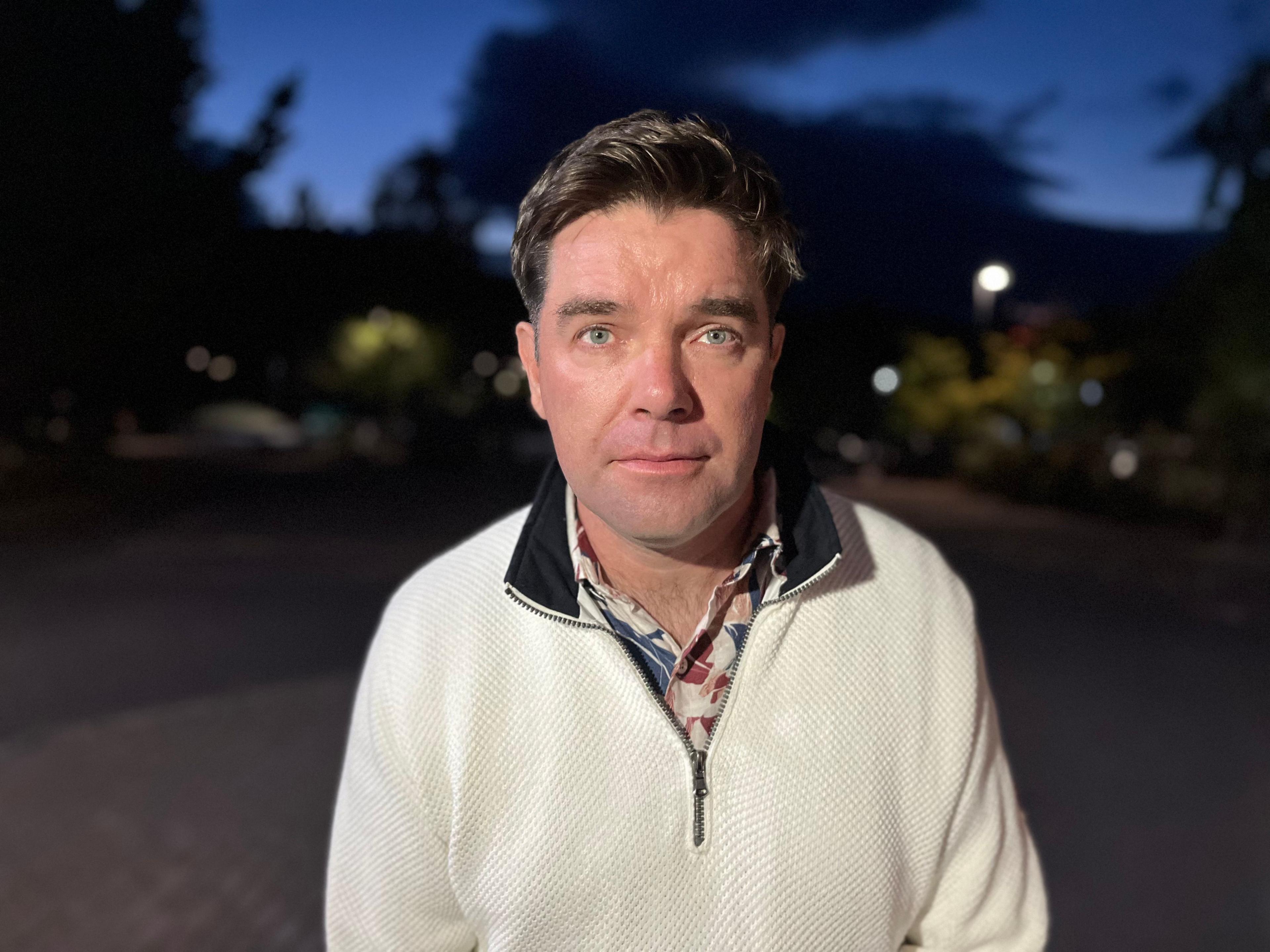 Local resident John McGlinchey wears a white quarter-zip sweater stands outdoors at dusk, facing the camera. The background is softly blurred, showing trees, lights and a darkening blue sky, with the subject illuminated by a nearby light source.
