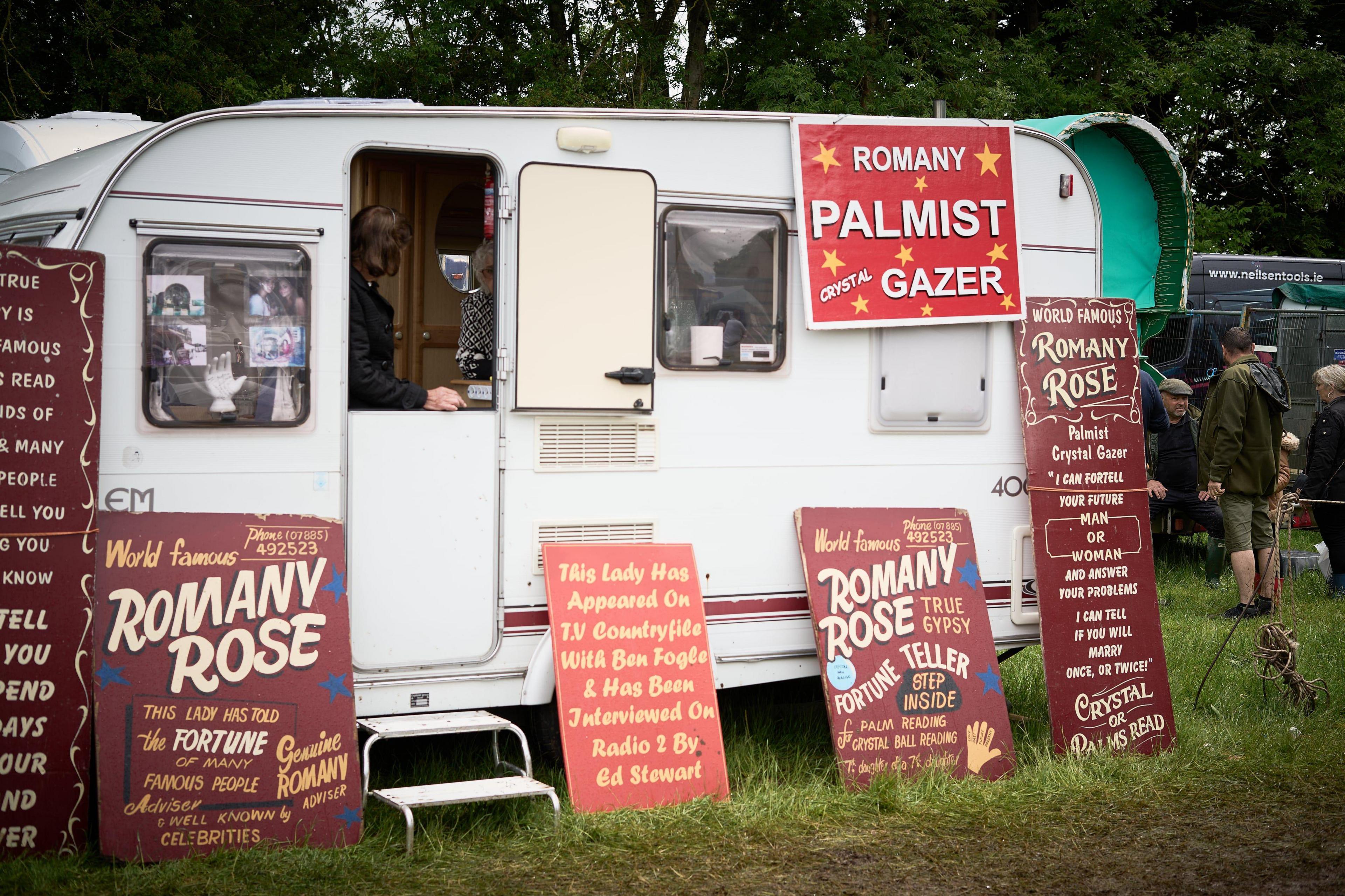 A caravan for the "Romany Palmist Crystal Gazer". The sign is in red and there are lots of other red signs attached to the white caravan.