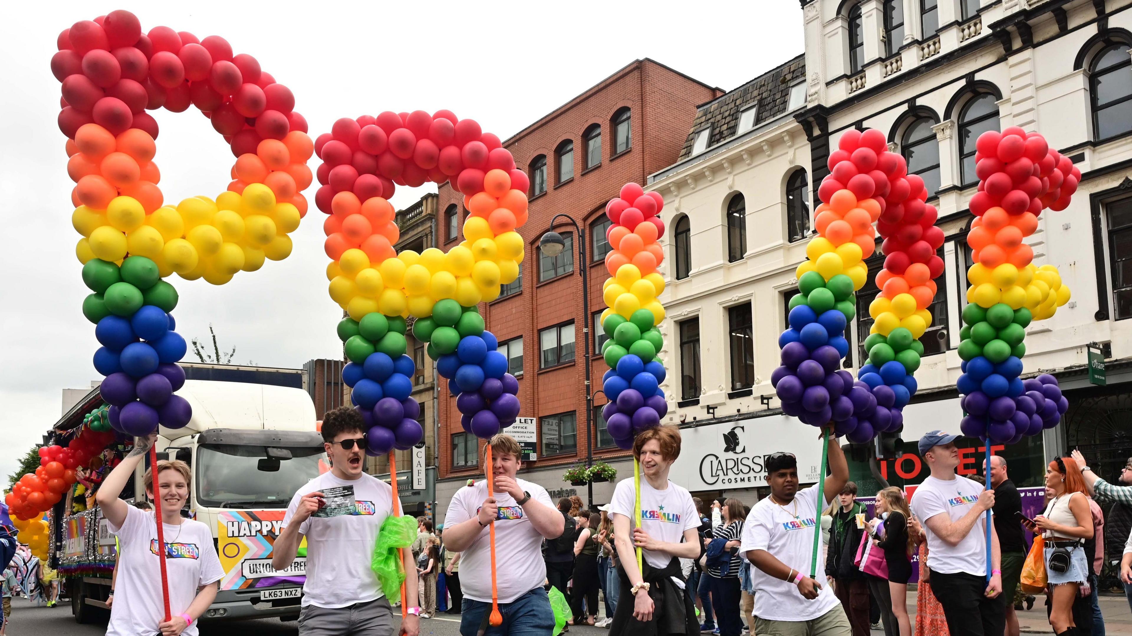 People taking part in the Belfast Pride parade in the city centre in 2024 on the street, they are wearing white t-shirts are holding balloons that read PRIDE in red, orange, yellow, green and blue. They are walking down a Belfast street with crowds watching on.