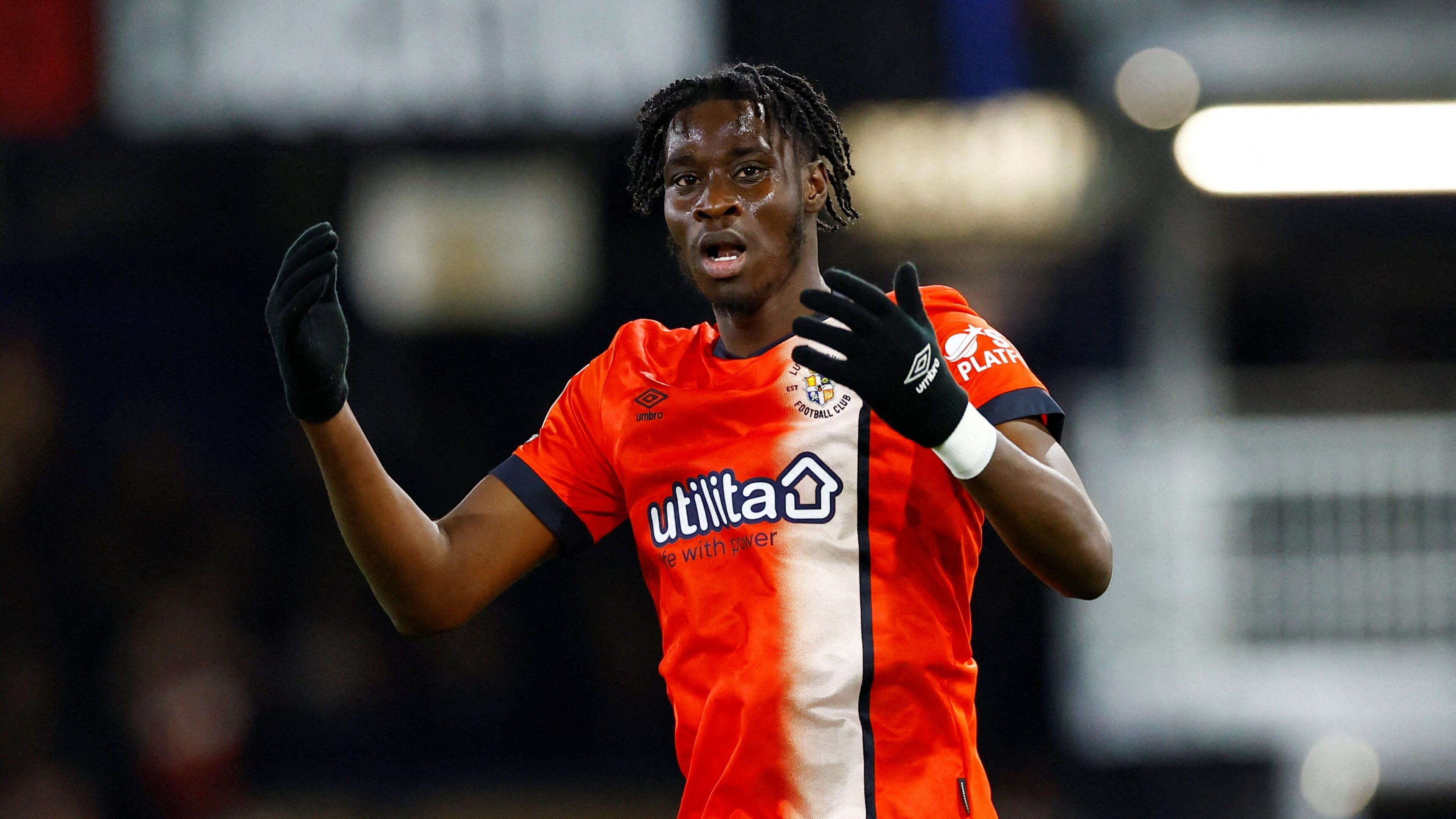 Elijah Adebayo has his mouth agape and throws his hands out in front of him in annoyance. He wears an orange luton football shirt and black gloves