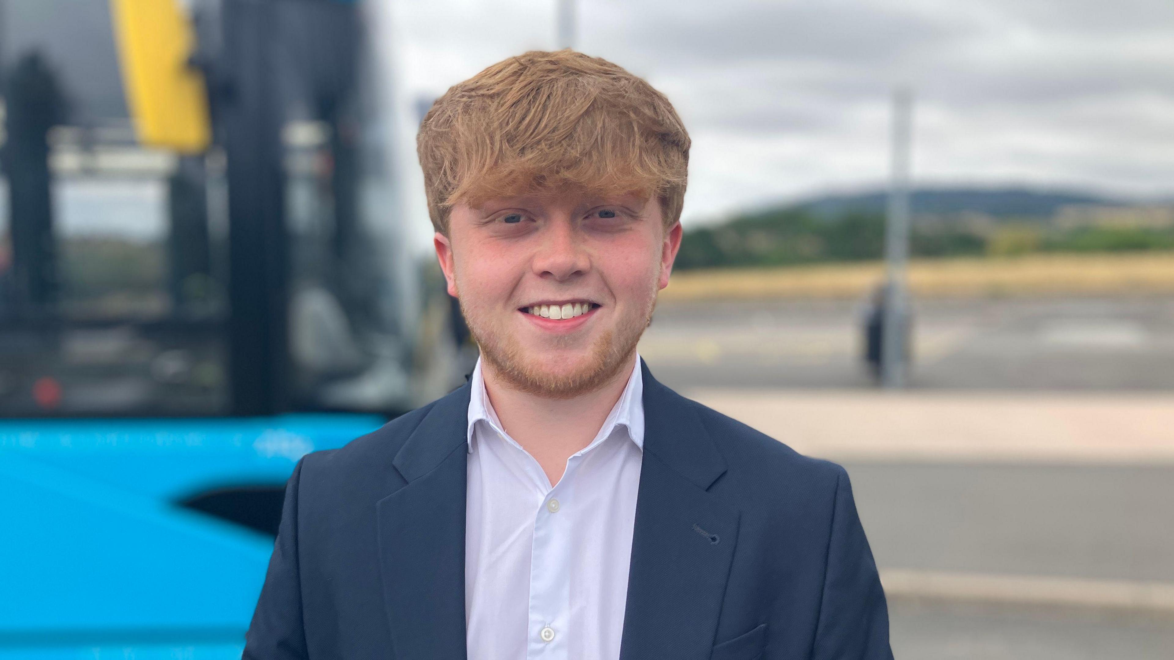 Ginger-haired man with a ginger beard, wearing a navy suit and white shirt, smiling in front of a blue single-decker bus, with a car park and hills in the background 