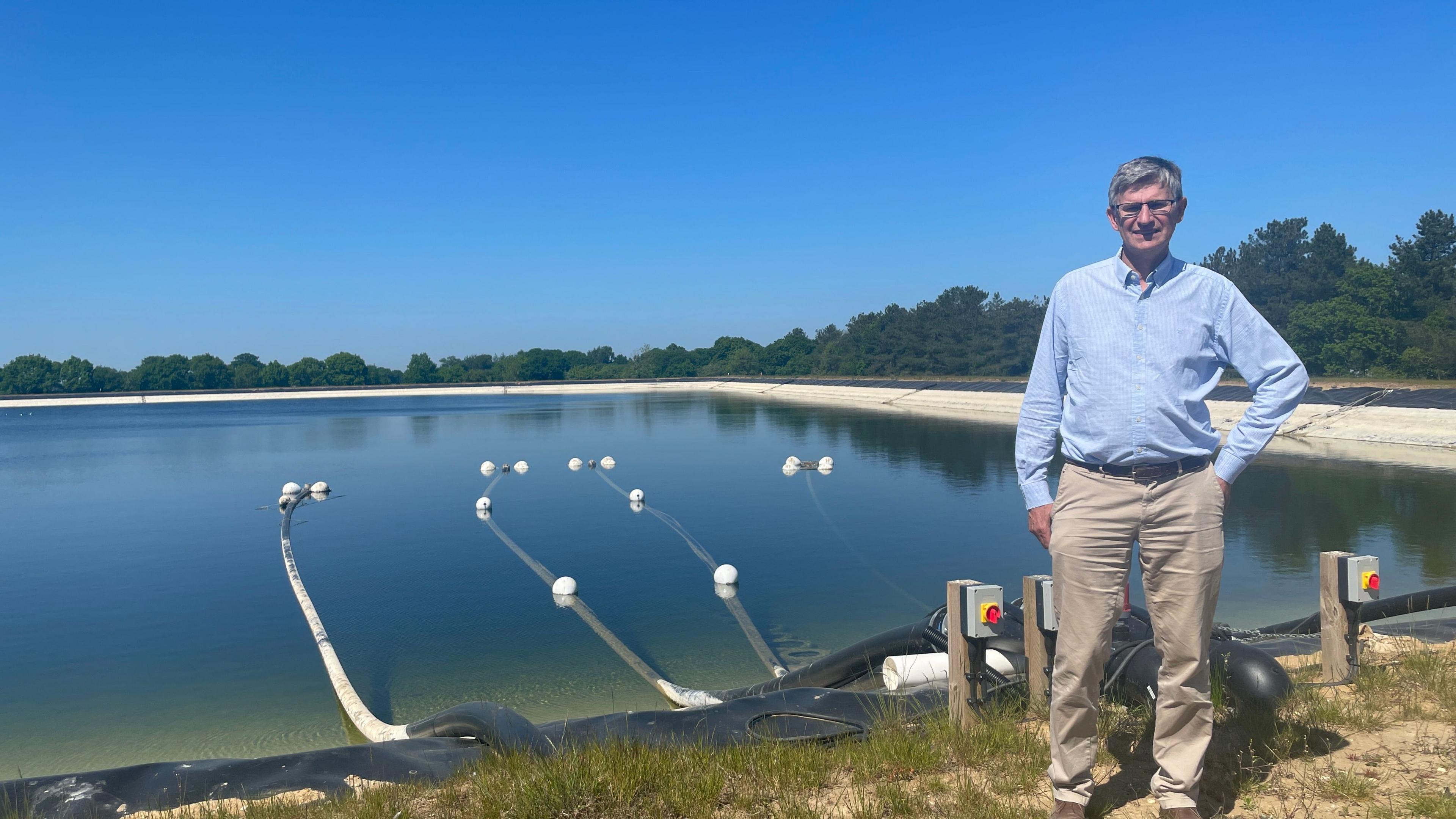 A man in a blue shirt and cream trousers stands next to a large reservoir with pipes leading out of it.