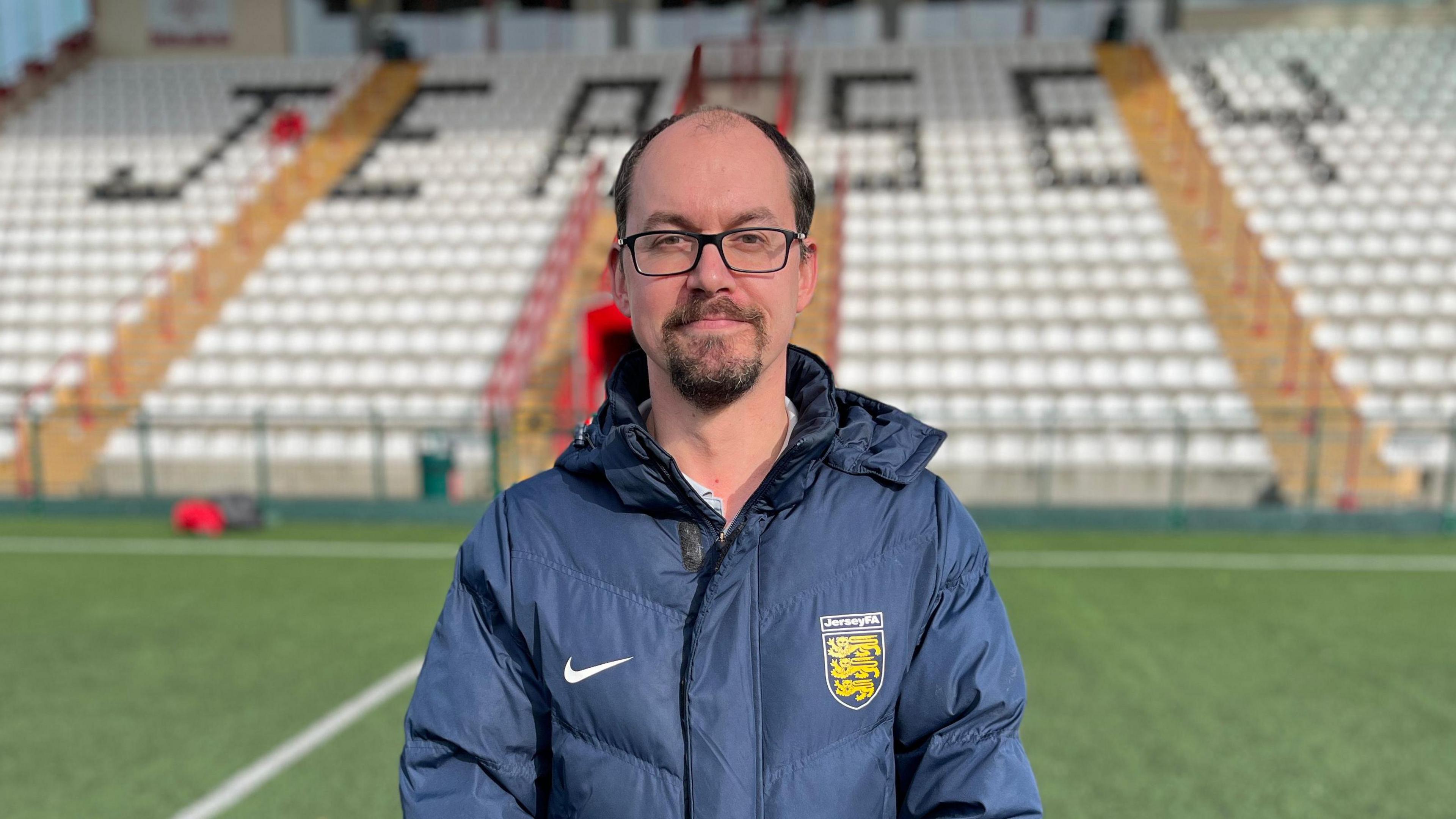 Tim Pryor wearing a navy blue coat with a Nike tick badge and a Jersey FA crest on the other side. He has a dark goatee beard, glasses and brown hair. Behind him is a football stadium's seats which are white with Jersey written out using black seats.