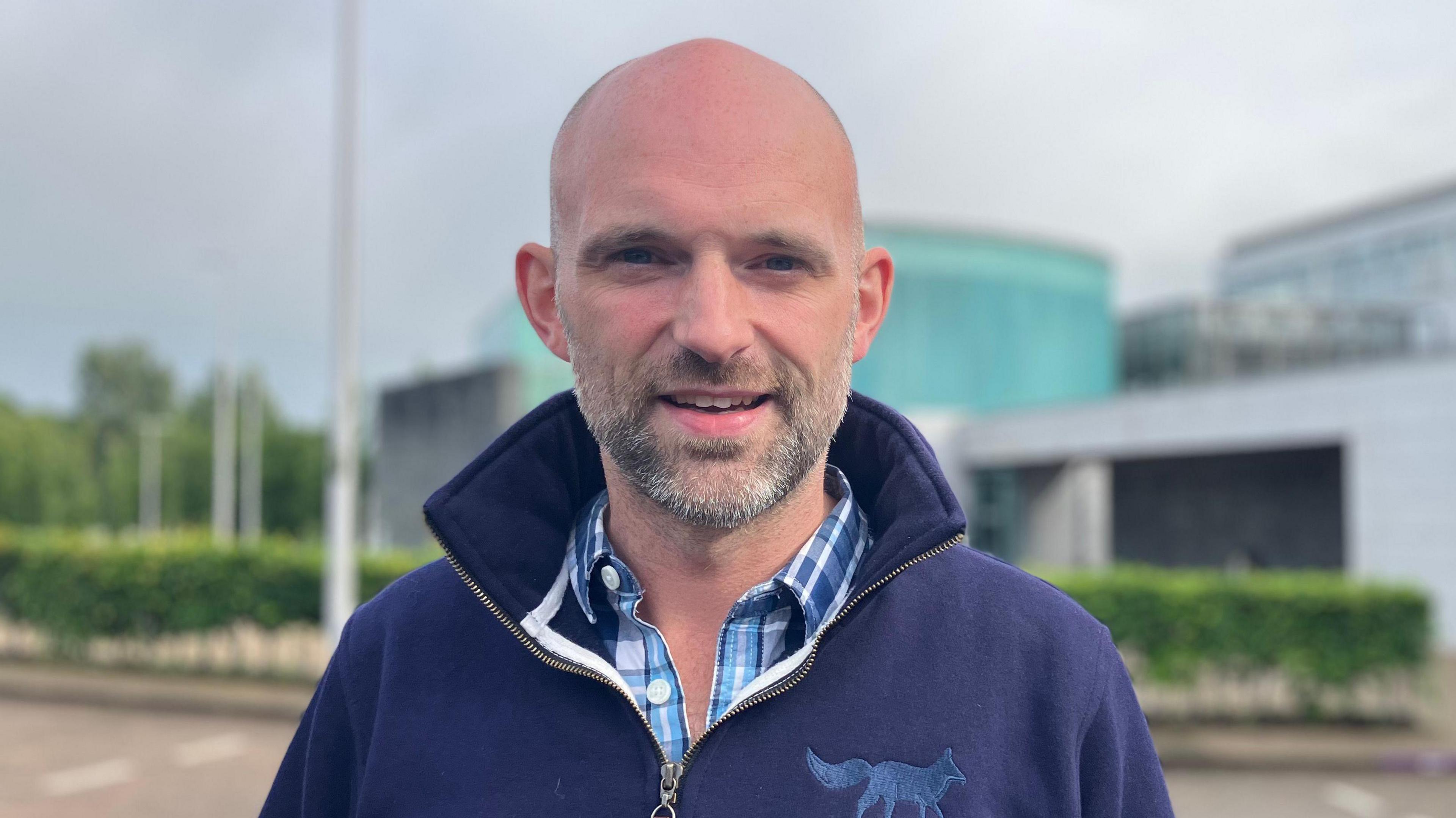 A man who is bald with a black and grey beard looks into the camera. He is smiling and is wearing a blue check shirt with a navy pullover. The pullover has the emblem of a fox on the front.
