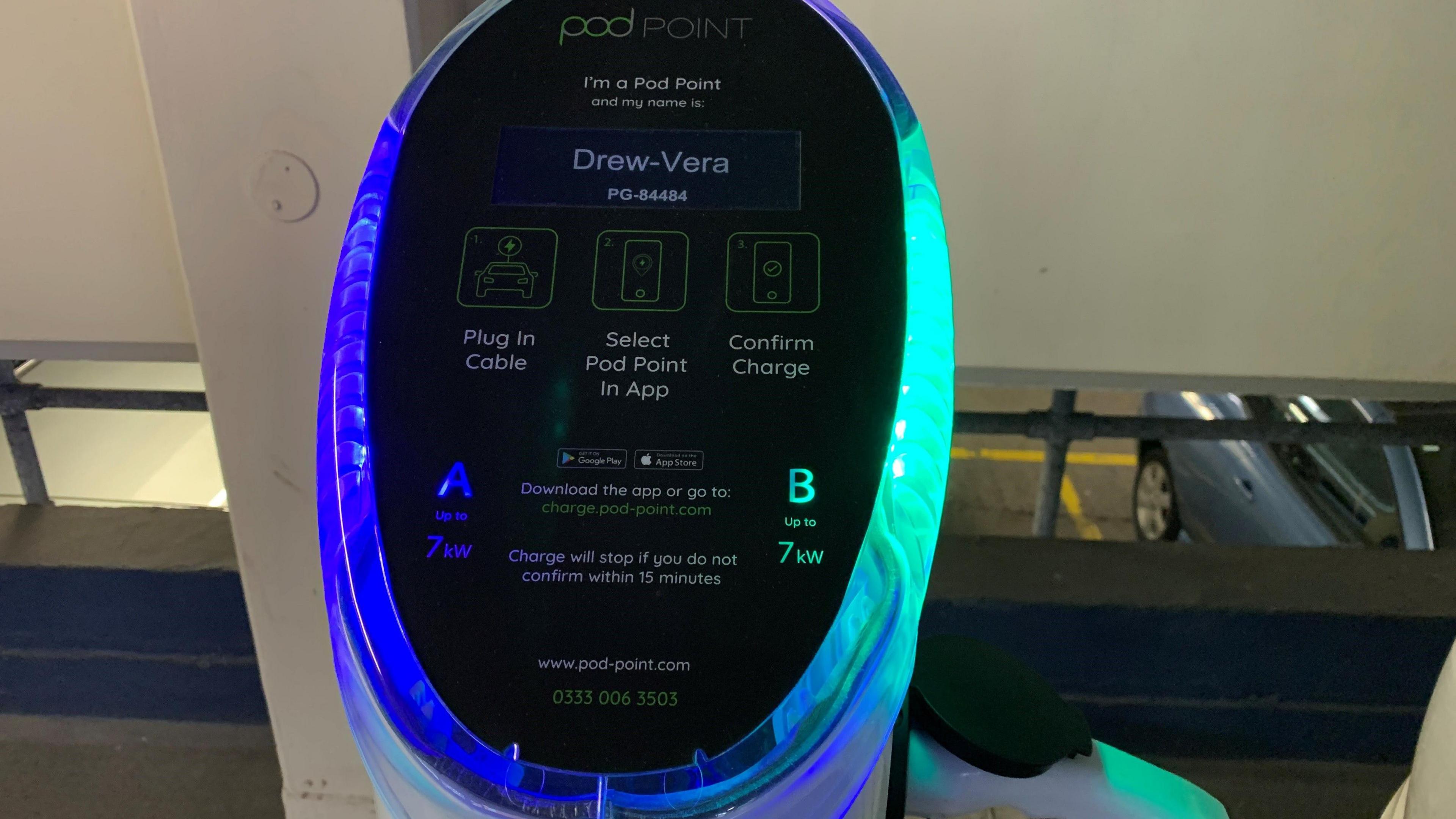 A closeup of the front panel of a 7kW Pod Point charging point in Chester Street Cart Park in Douglas. It has a black screen featuring instructions for using the charging point and blue and green light around the circular panel indicating it is charging a vehicle.