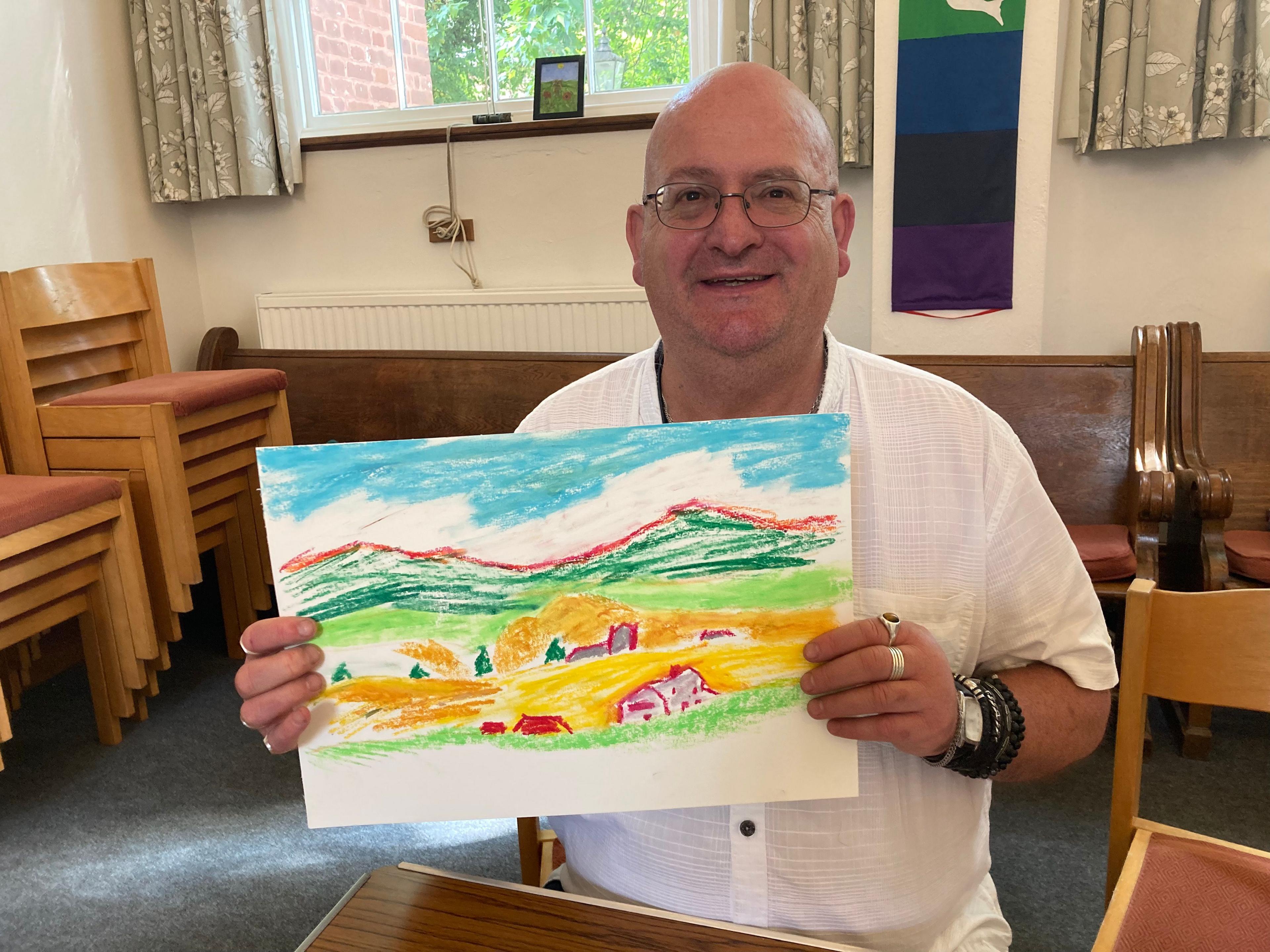 A man wearing a white shirt holds up a picture depicting a rural scene with hills in the background and buildings in the foreground. The man is sat down, and a window with a set of patterned curtains can be seen behind him as well as stacks of chairs and a radiator behind a wooden bench.