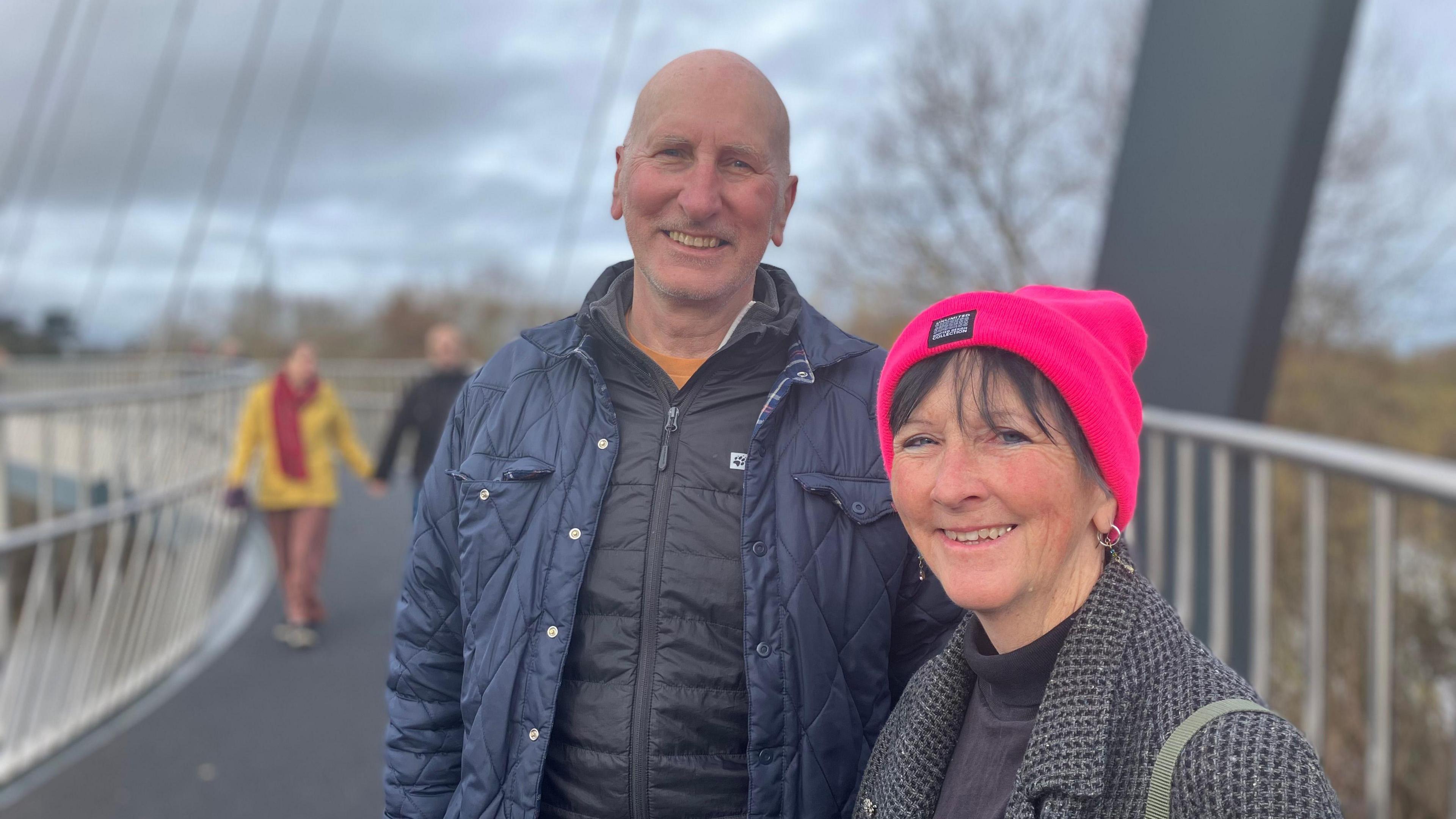 A bald man smiling and wearing a blue quilted jacket with a zipped up black jacket underneath. He is standing next to a woman with a bright pink hat, grey patterned coat and black turtle neck top. She is also smiling while they both stand on the Kepax Bridge