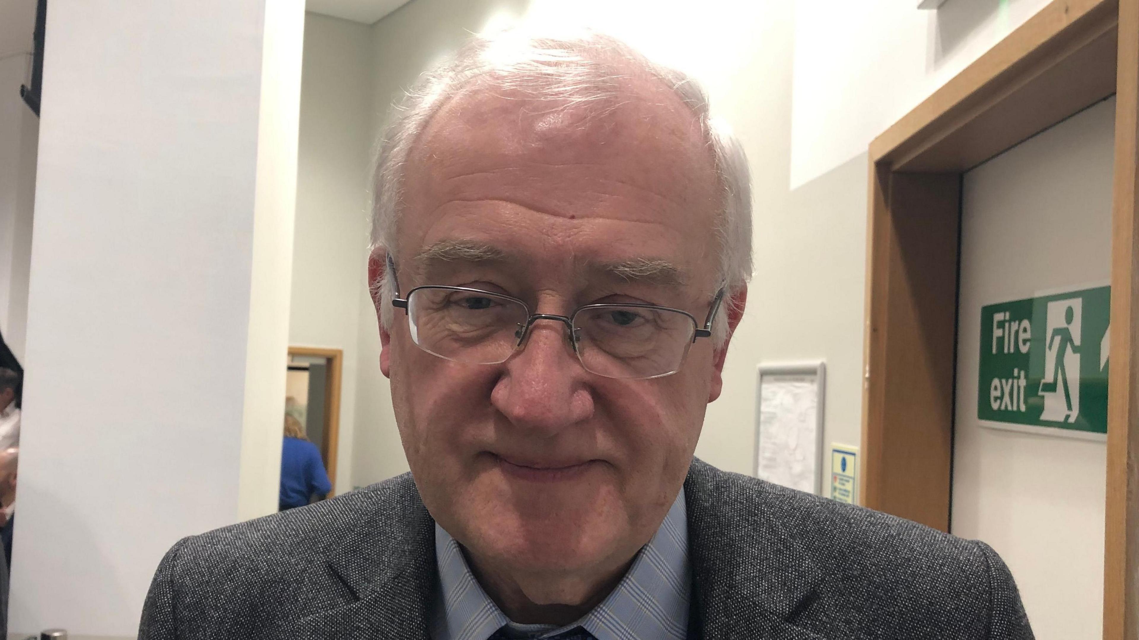 Christopher Hudson smiles at the camera. He has grey hair and glasses. He wears a grey suit with a light blue shirt and blue tie.