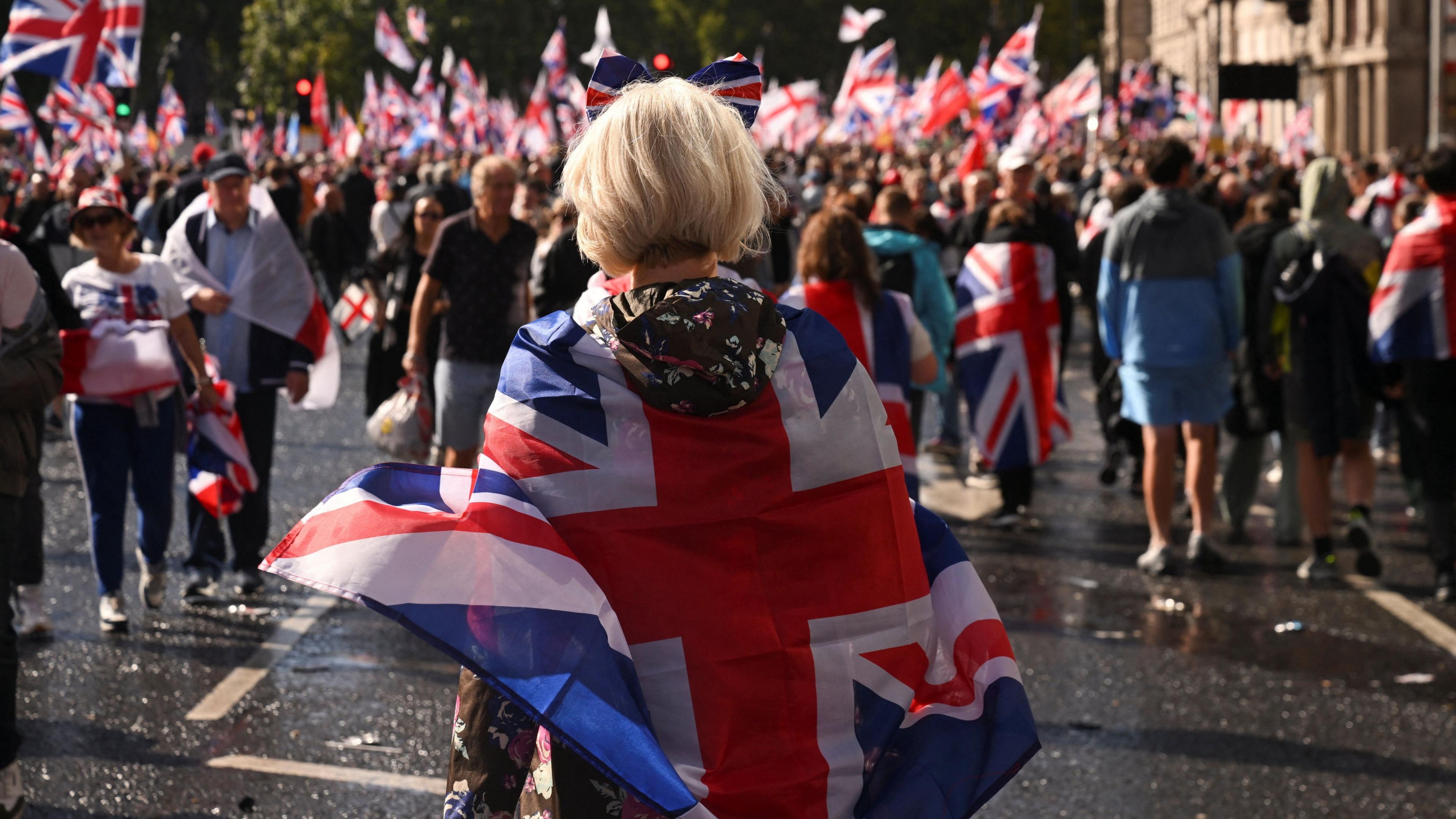 A woman wearing a Union Jack flag draped over her attends a rally with people carrying flags in the crowd in front of her
