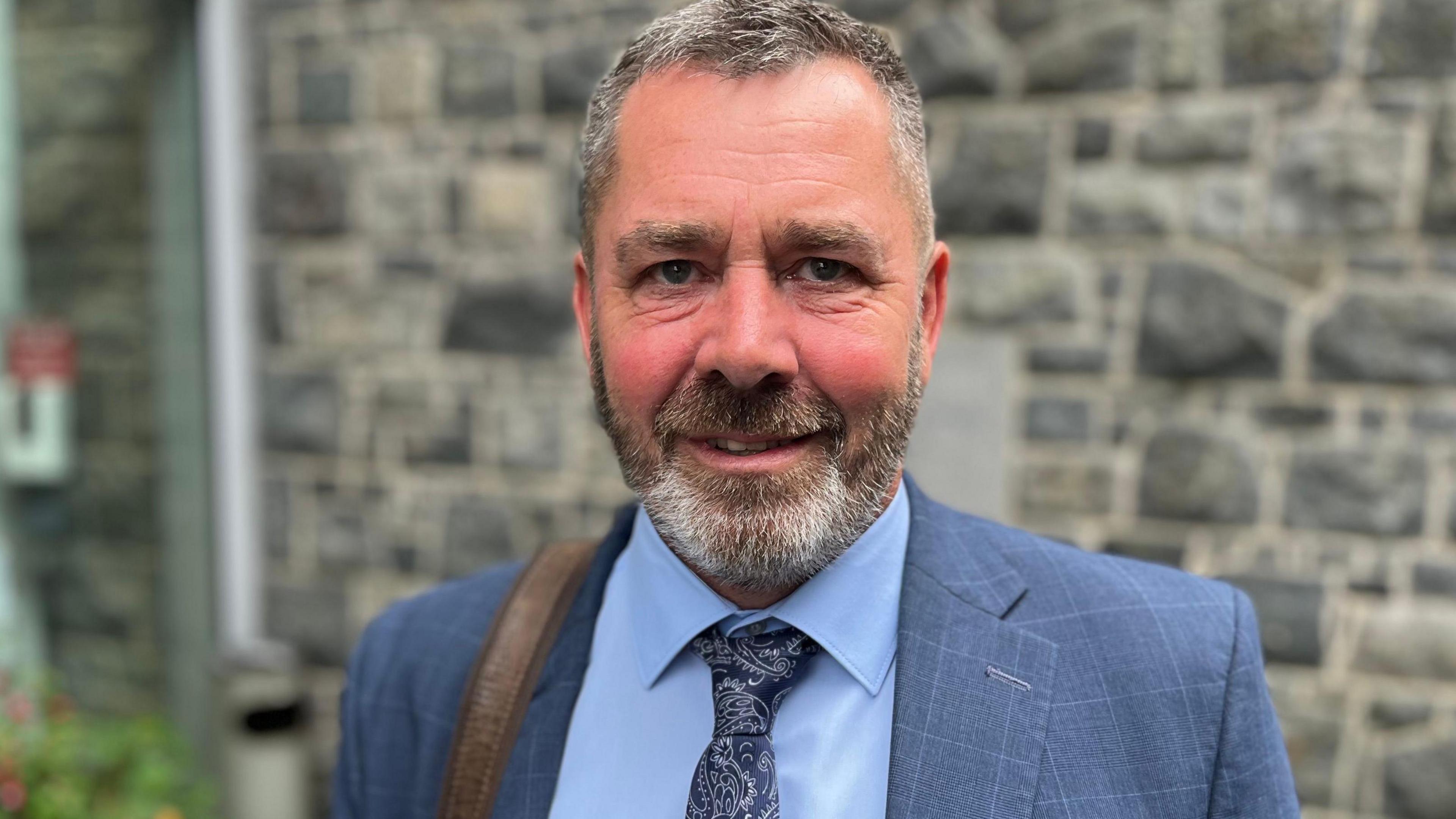 Deputy Marc Leadbeater - A man with grey and black hair, with a grey and black beard looking at the camera. 
He is wearing a blue suit jacket, blue shirt and blue patterned tie.