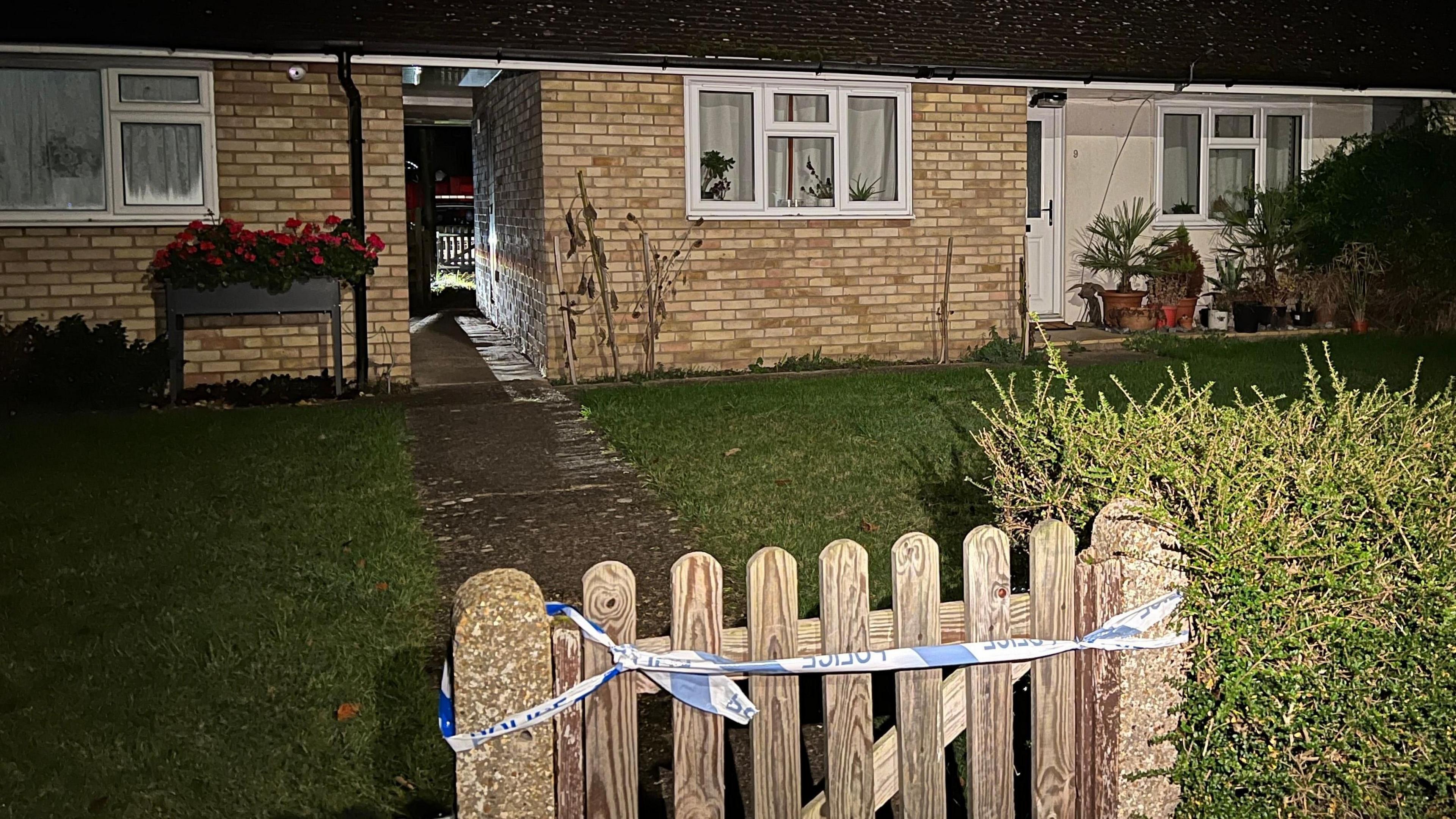 The front garden of a bungalow consisting of a lawn and a path leading to the building's front door. It is dark. A piece of police tape has been tied to a wooden gate leading to the path.
