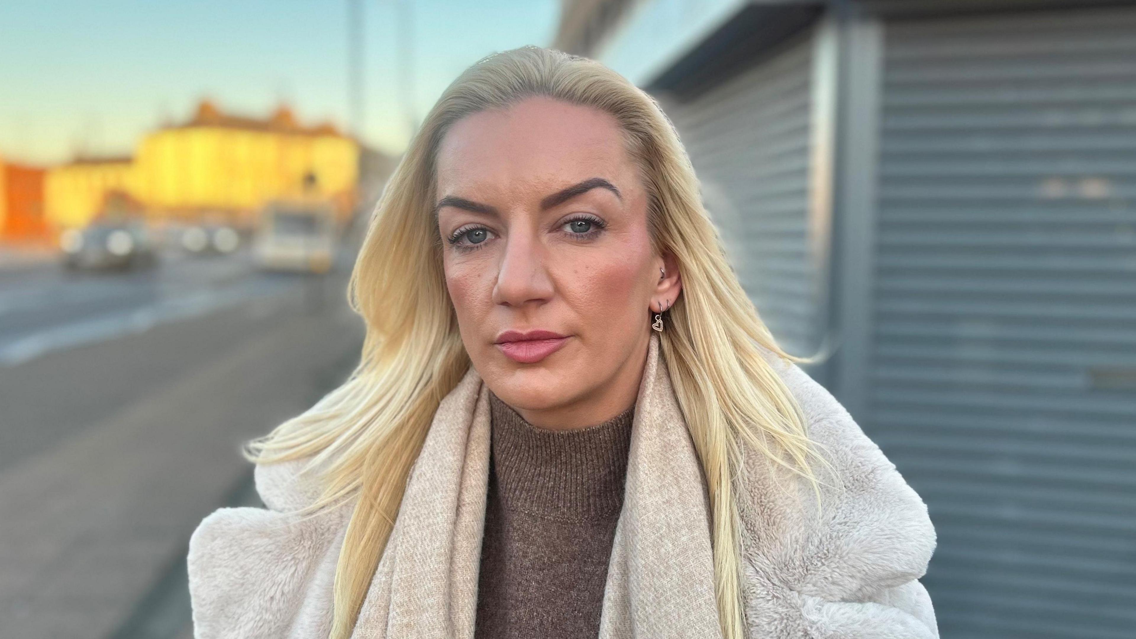 Amy, who has long blonde hair, stares into the camera while standing in a street. The grey metal shutters of a closed shop can be seen behind her. She is wearing a thick, off-white winter coat over a brown, high-neck jumper.