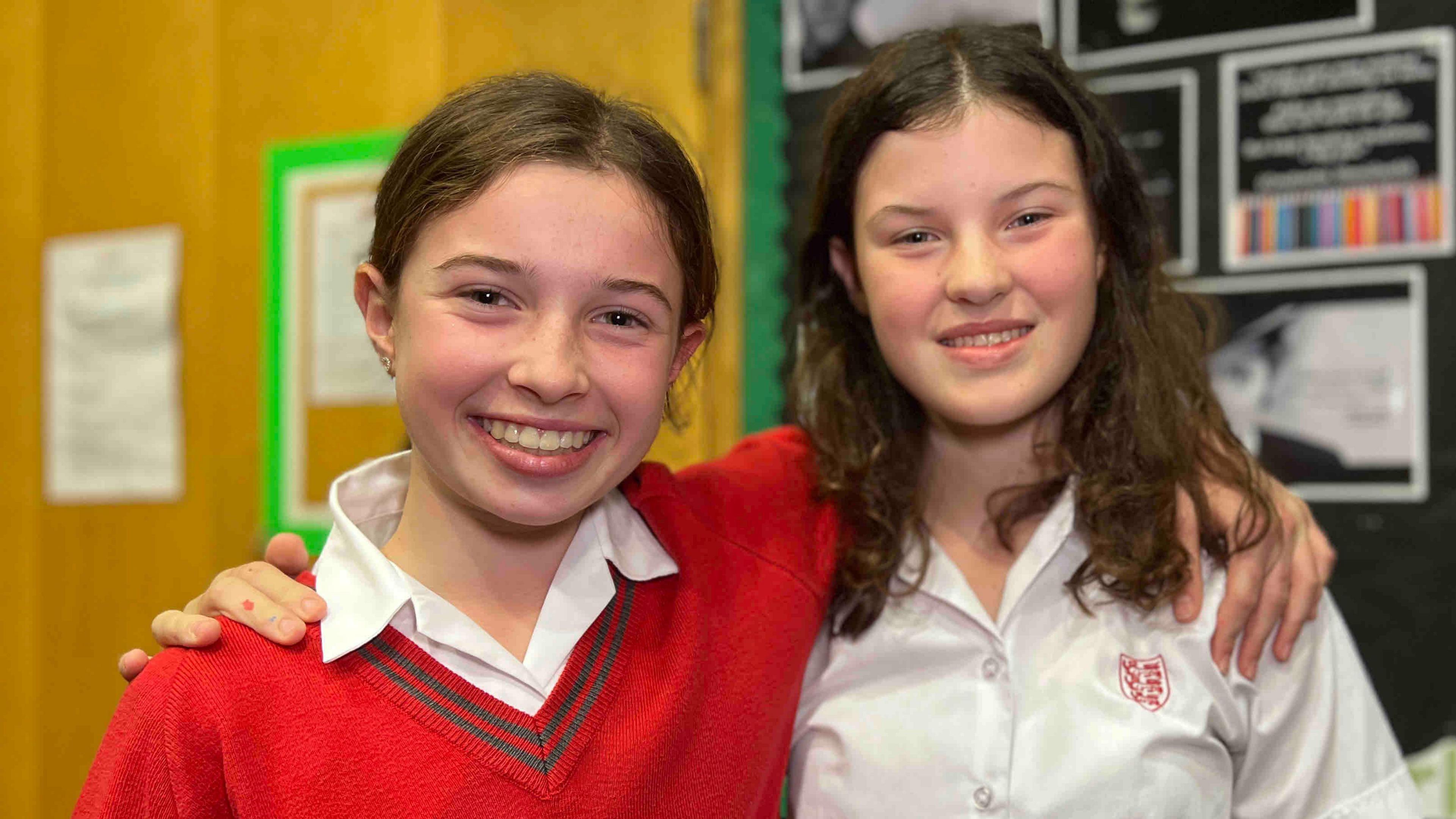 Florence and Esther are standing side by side in a classroom or rehearsal space. One is wearing a bright red sweater over a white collared shirt and the other is wearing a white button-up shirt with a small crest on the chest.