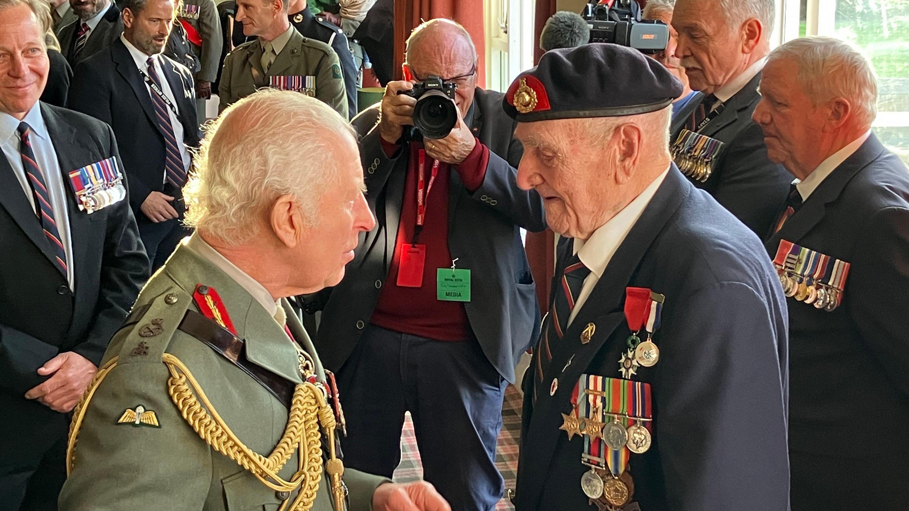 King Charles III speaks to 100-year-old D-Day veteran Norman Ashford, who is wearing a blazer with his military medals on, while surrounded by other former Royal Marines. 