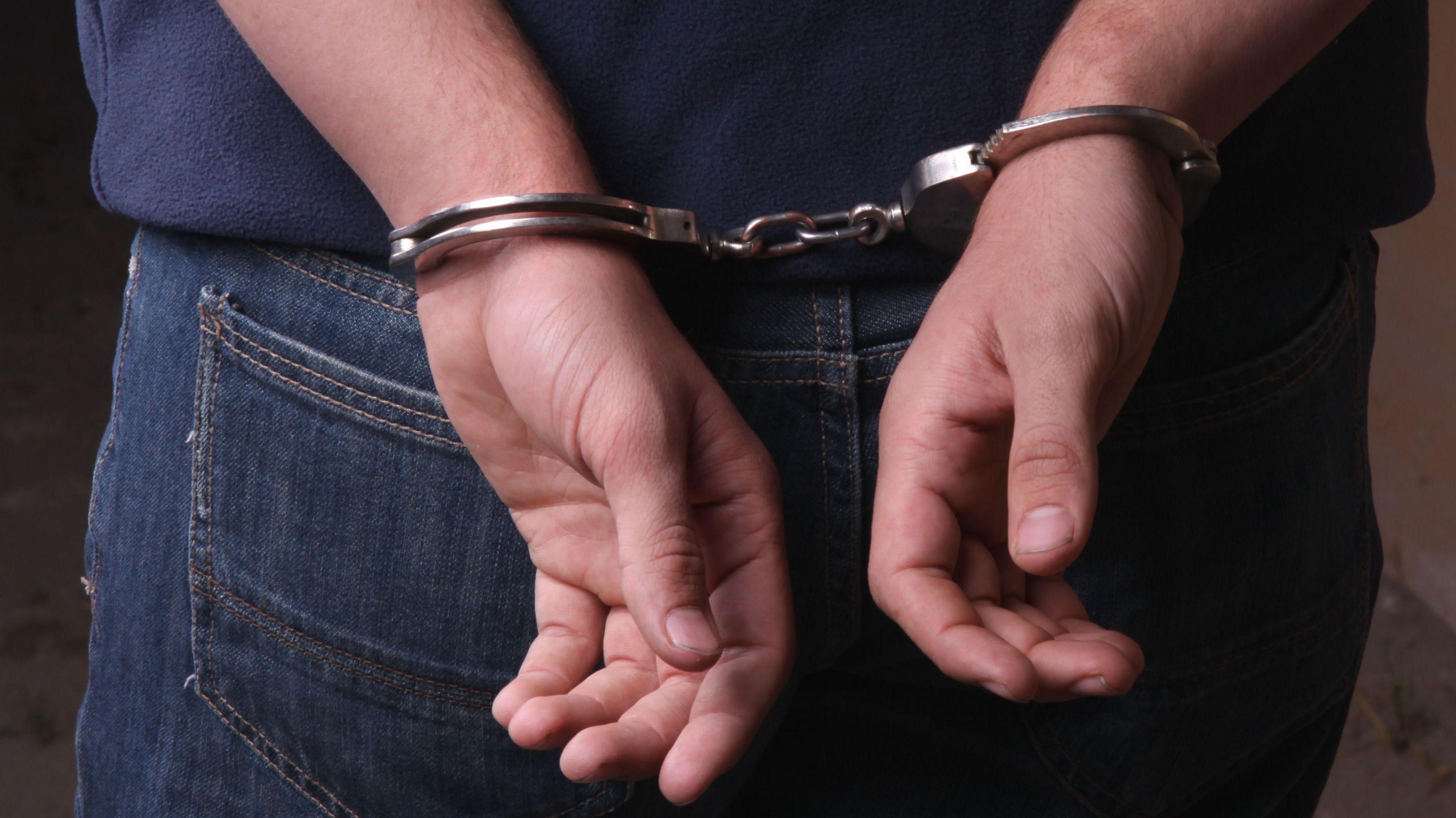 A close up image of a man's back with his hands cuffed following an arrest. He is wearing blue jeans and a dark blue sweatshirt.