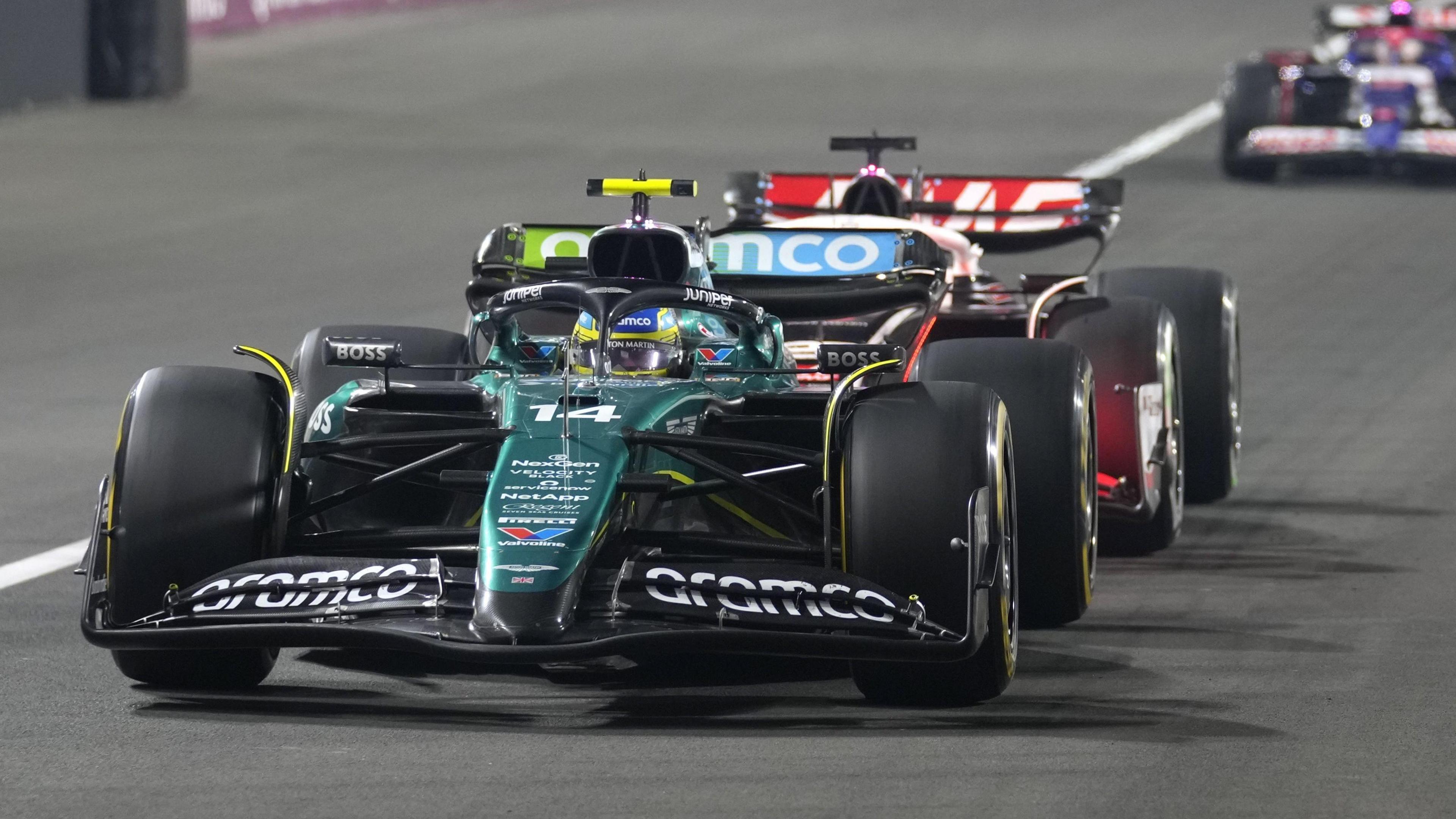 A green racing car with large shiny wheels driving forwards on the track with other cars behind it.