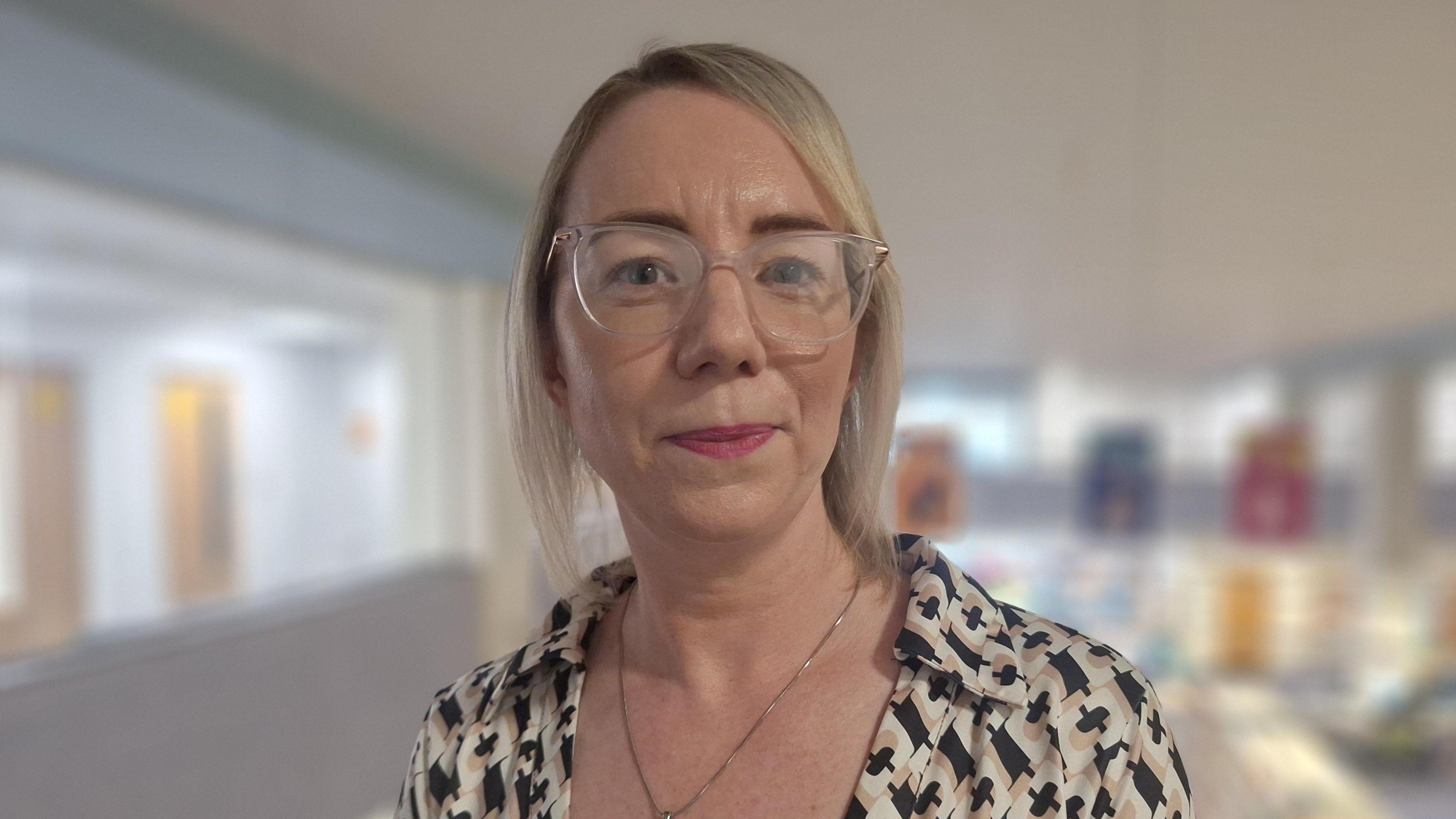 Kath Howells has blonde straight hair cut in a long bob. She's looking into the camera with the backdrop of the interior of Newport Central Library behind her. She wears glasses with clear frames, silver chain and a black, white and beige wraparound dress.