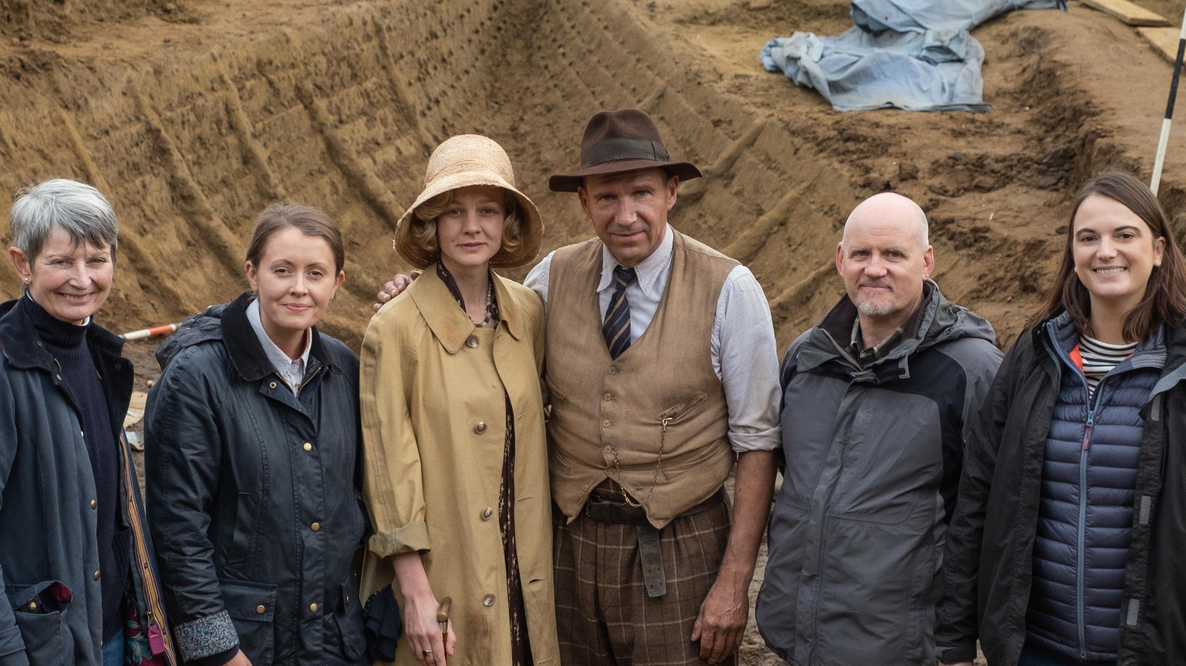 Members of Sutton Hoo, Netflix crew members and actors Carey Mulligan and Ralph Fiennes, stand in a line and smile at the camera. Behind them is a fake excavation site.