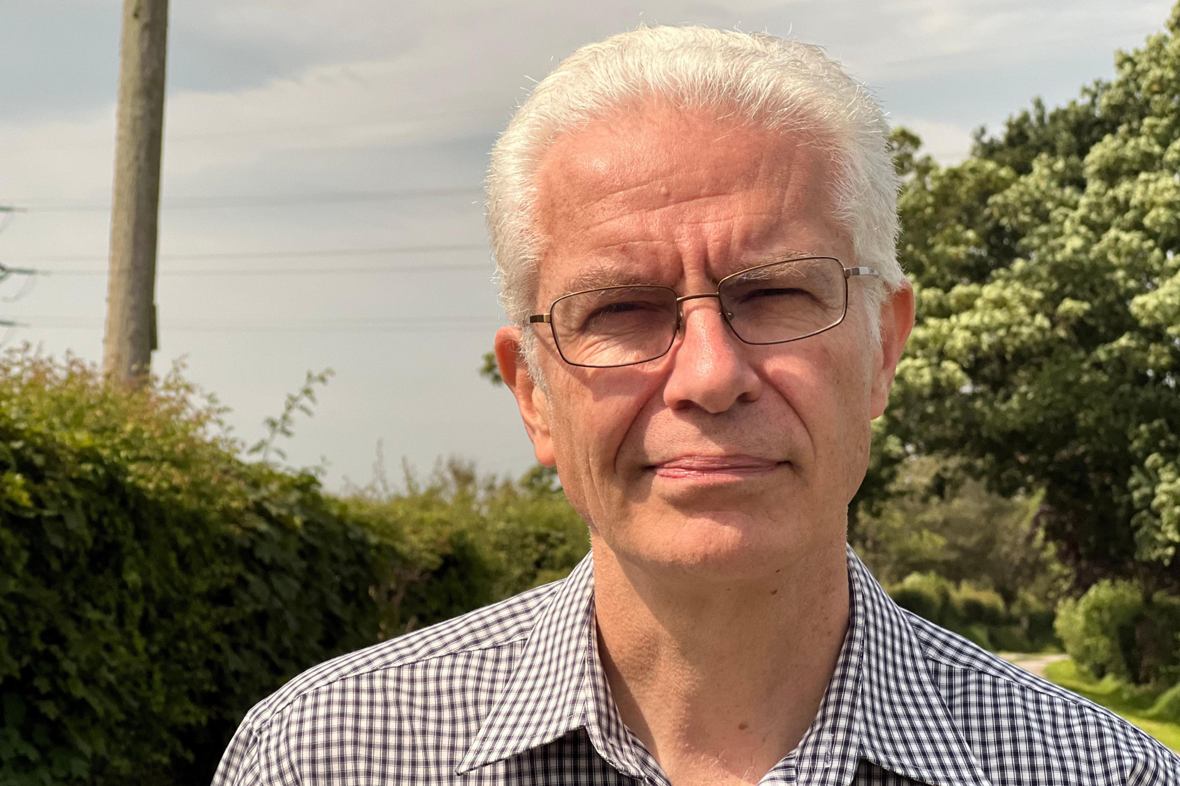 David Mudge is wearing glasses and a check patterned shirt, and has short, white hair. He is clean shaved. There are trees and hedgerows in the background.