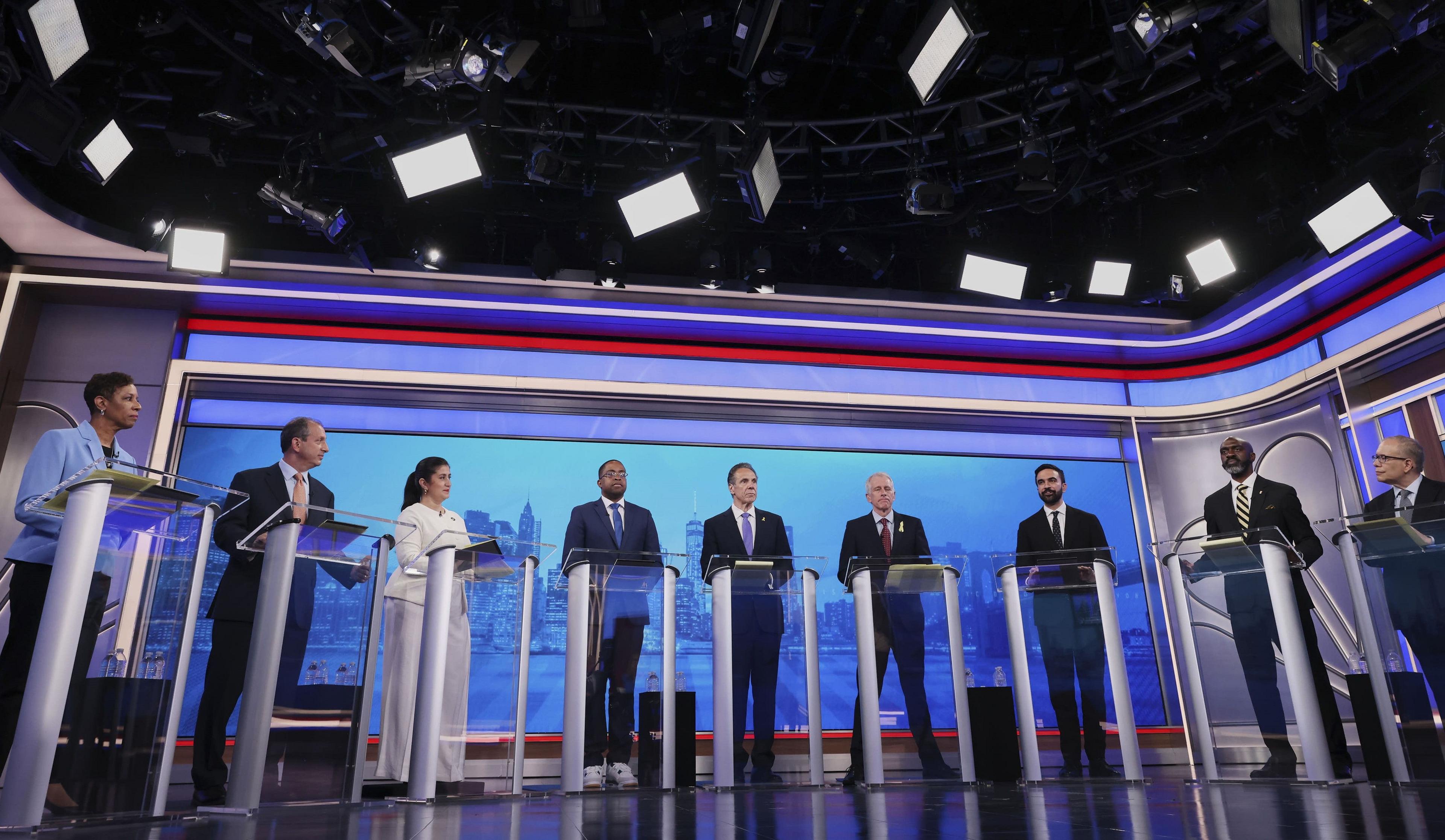 Nine candidates stand at podiums on a stage, each behind a glass podium.