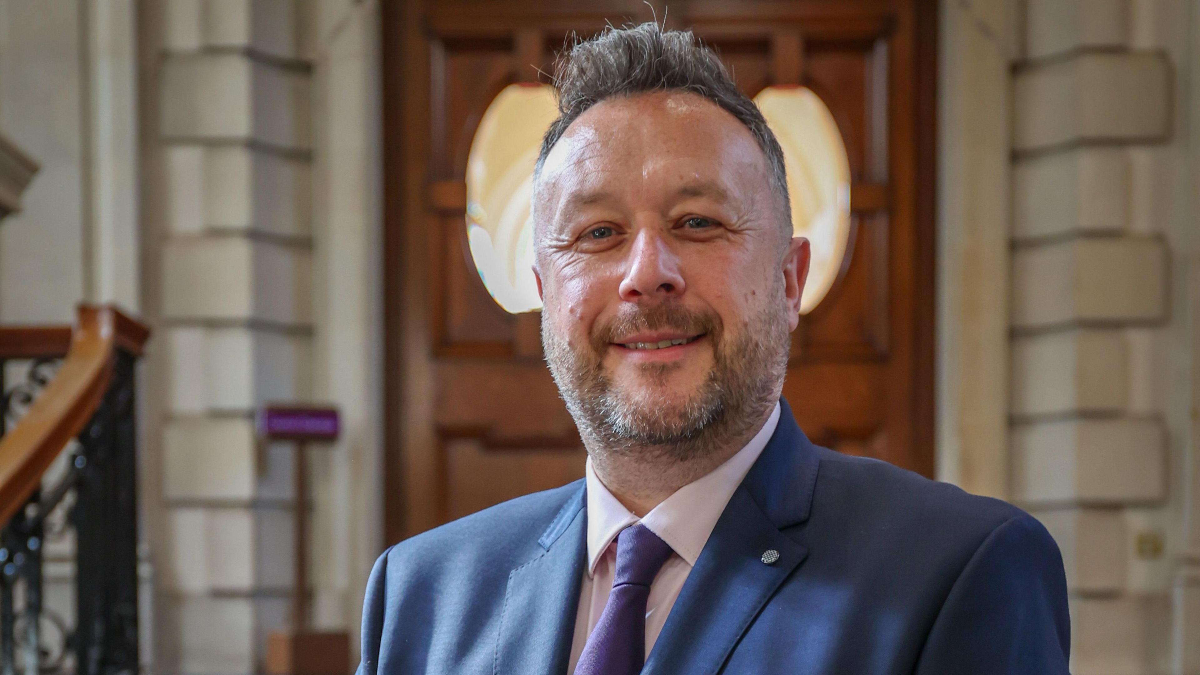 A mean with short grey hair and a grey beard is wearing a blue suit jacket, blue tie and white shift standing in front of a wooden door and stairs.