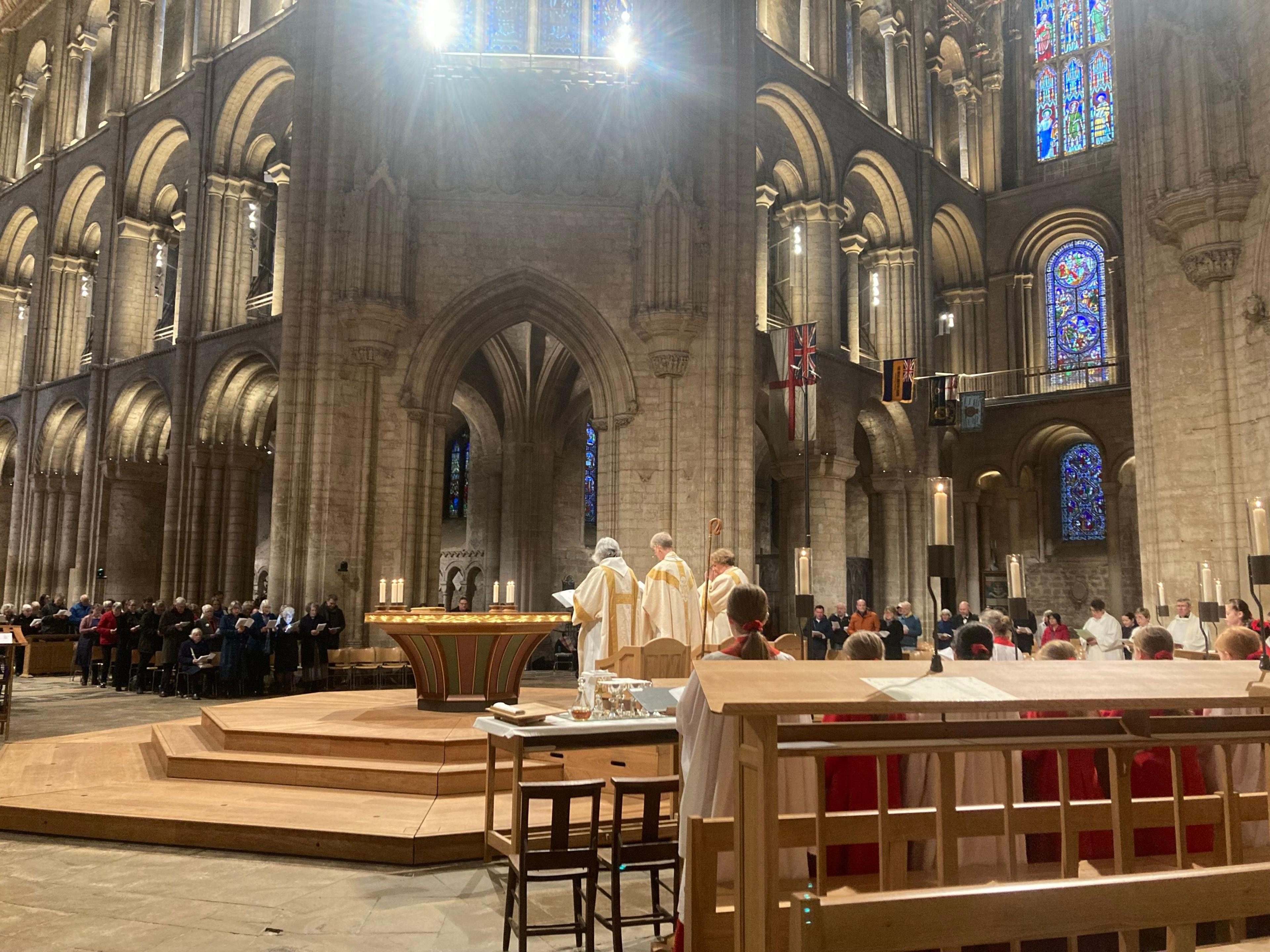 Ely Cathedral seen from inside looking towards the congregation as a service is held. Three priests in white and gold robes stand at the front, leading the service, while a choir stands behind them, seen from behind. here are candles and blue stained glass windows.