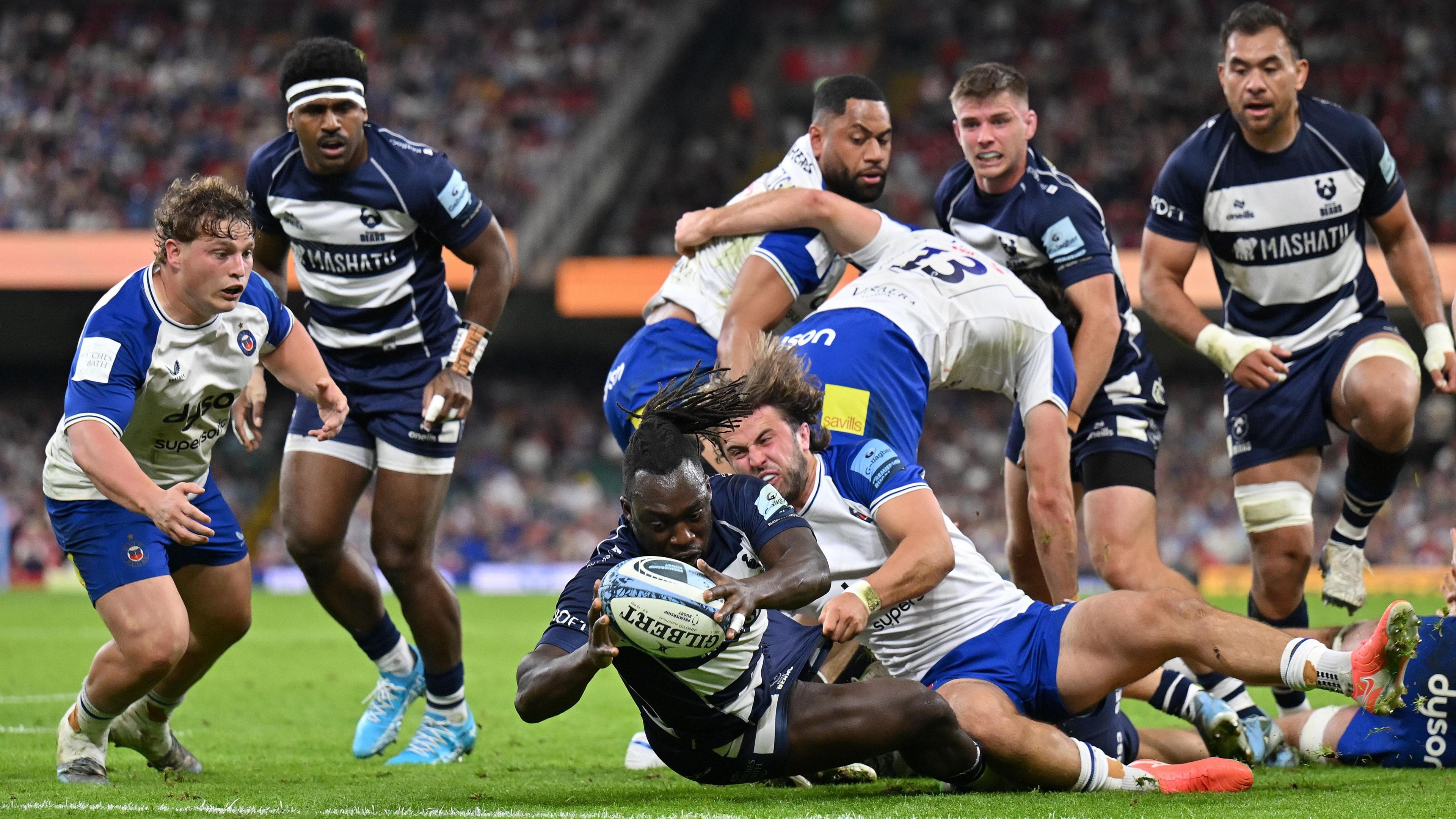 Bath Rugby and Bristol Bears players in a tackle. There are eight male players in the shot. One Bristol player has fallen to the ground and is holding the ball, while his opponent from Bath is trying to grab it from him. The crowd can be seen in the distance.