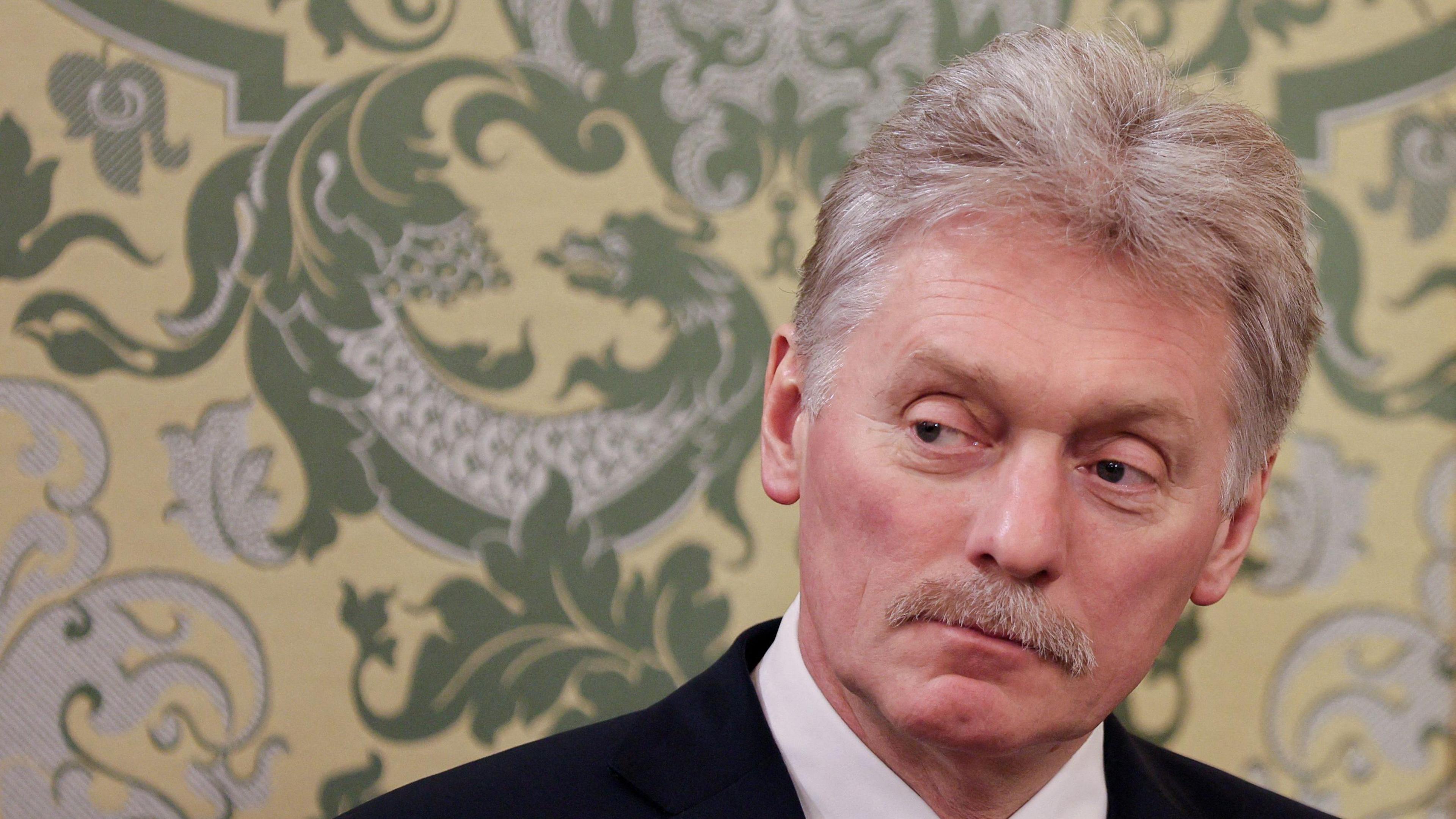 Headshot of Dmitry Peskov, who has white-grey hair and a moustache. A yellow wallpaper with a green dragon print is on the wall behind him.